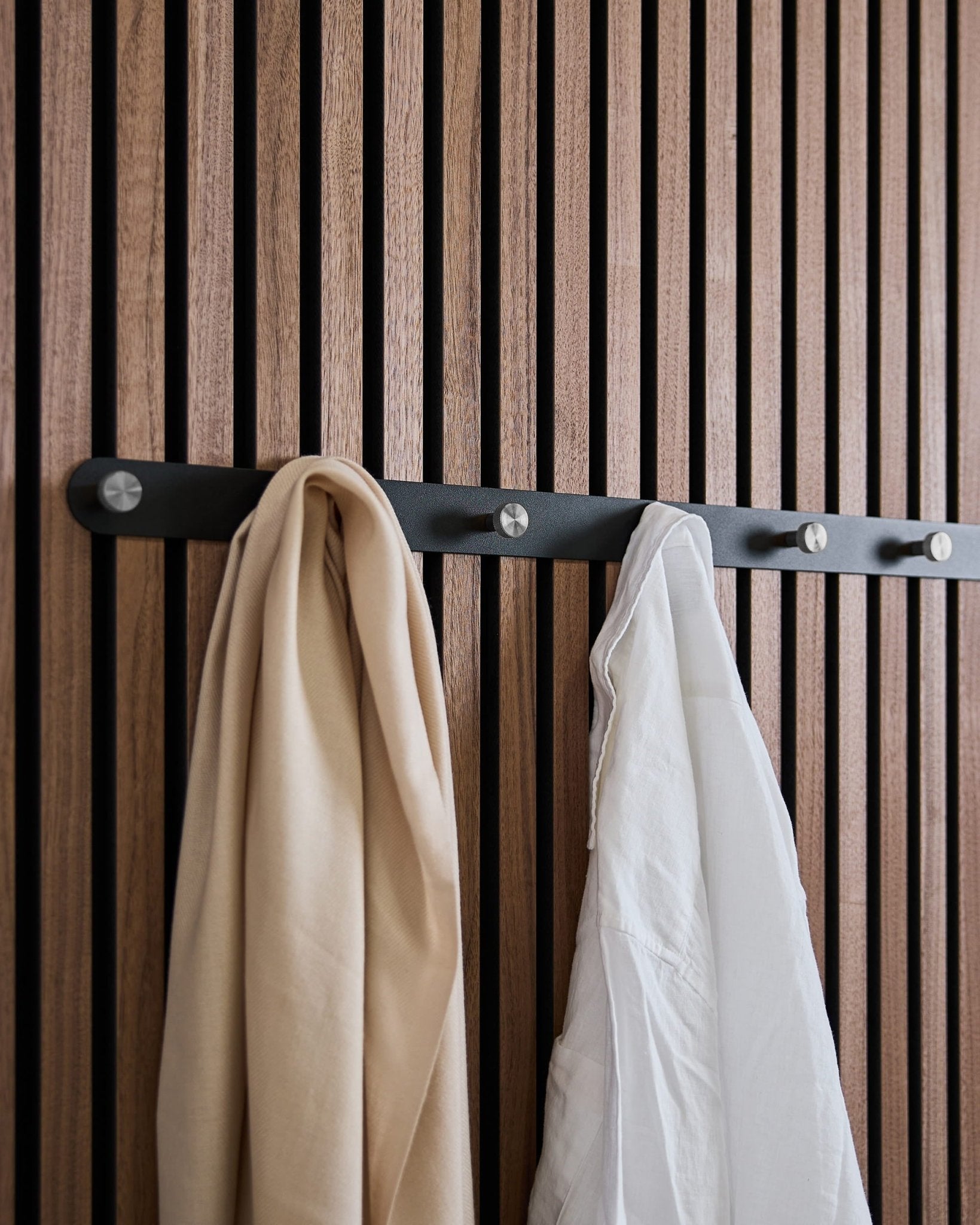 Et beige tørklæde og en hvid skjorte hænger på WoodUpps Norr X7 Black/Stainless Steel knagerække, der er monteret på et nordisk akustisk lamelvægspanel.