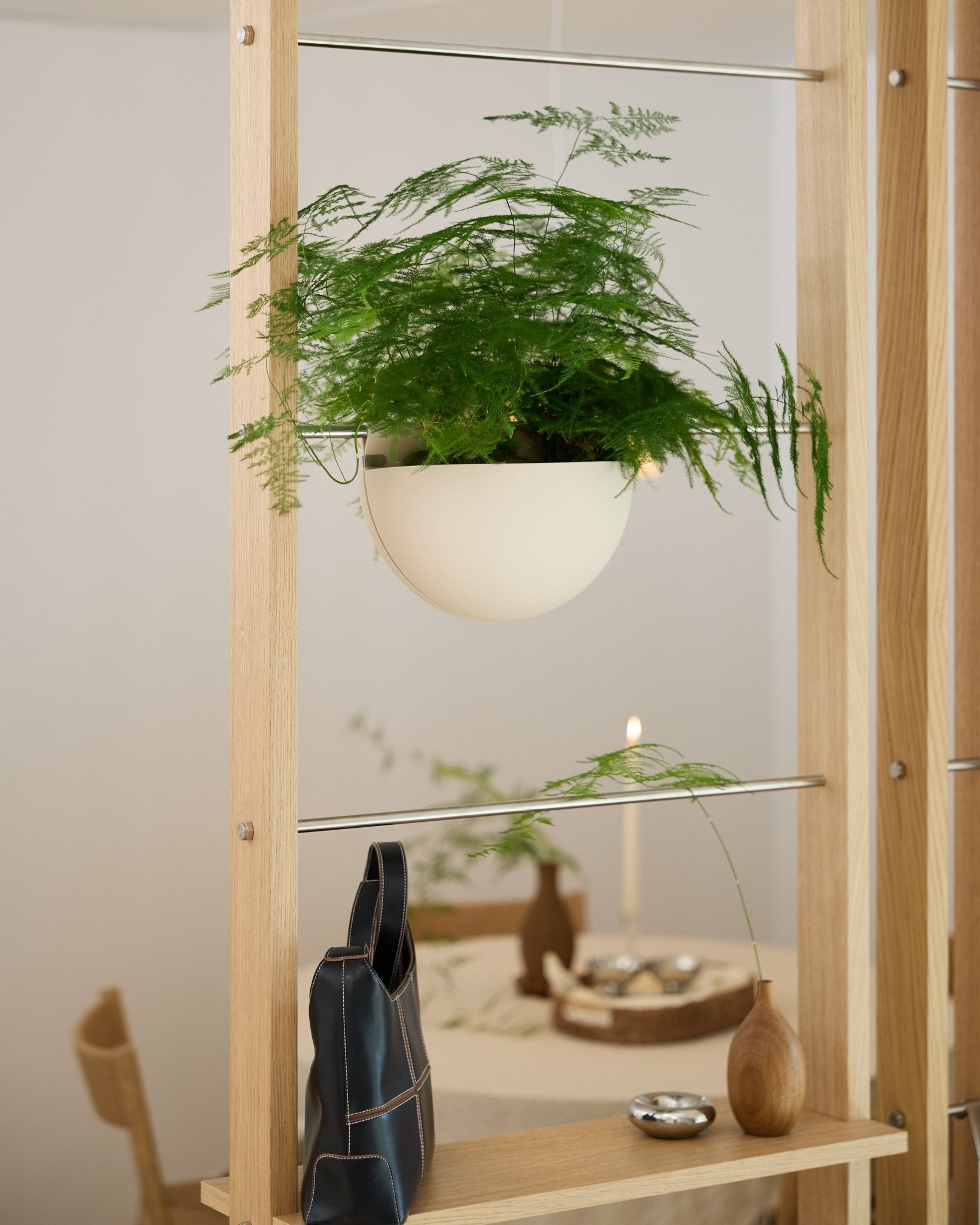 En grøn bregne i en lysbeige WoodUpp Gro Soft Pocket BM2-plantekrukke står på en træhylde over en sort taske, en vase og et dækket spisebord.