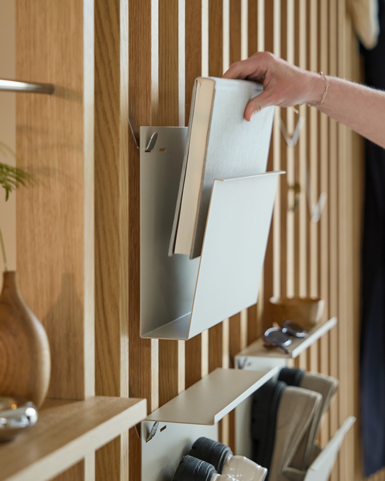 En hånd placerer Nell | Magazine Storage / Beige af WoodUpp på en pulverlakeret stålhylde monteret på et nordisk akustikpanel af træ med ting i nærheden.