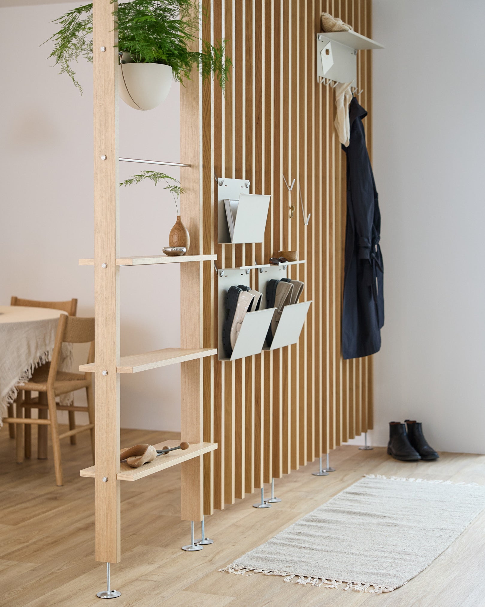 WoodUpp Nell Magazine Storage i beige integreret i en rumdeler med stålhylder, opbevaringskasser og knager; spiseplads og tæppe er synlige bagved.