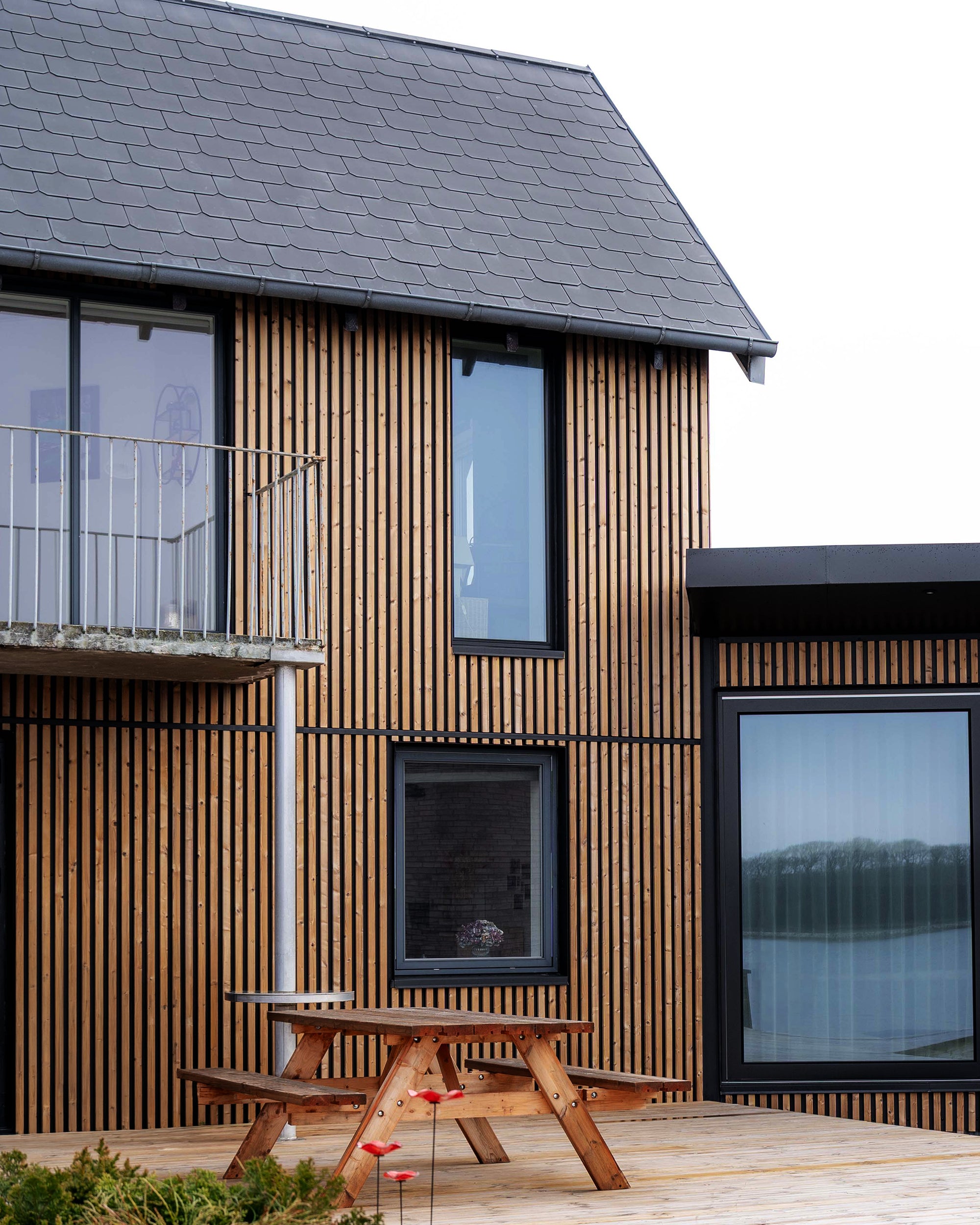 Modern house featuring vertical Aluwood facade panels, large windows, and a small balcony; a wooden picnic table on the patio adjacent to the garden.