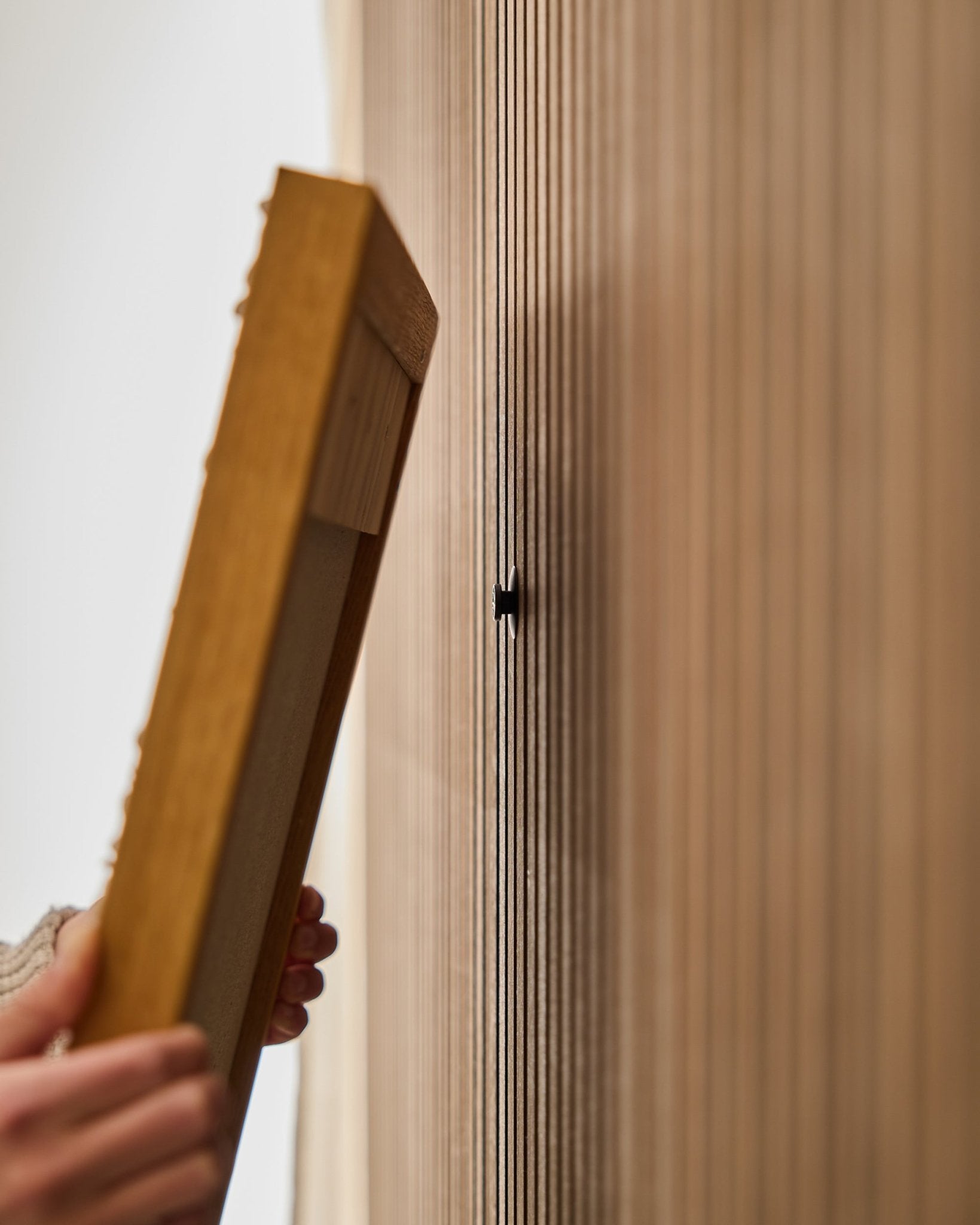 En person bruger WoodUpp Dupps | Original-sættet til at montere en Akupanel ramme til akustisk træpanel med fokus på at fastgøre en small black krog tæt ved vertical lameller.