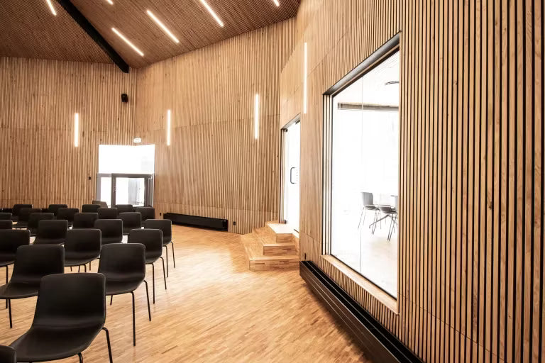 Rummeligt konferencelokale med Akupanel akustiske træpaneler på vægge og loft, rækker af sorte stole, store vinduer og lodrette lysarmaturer.