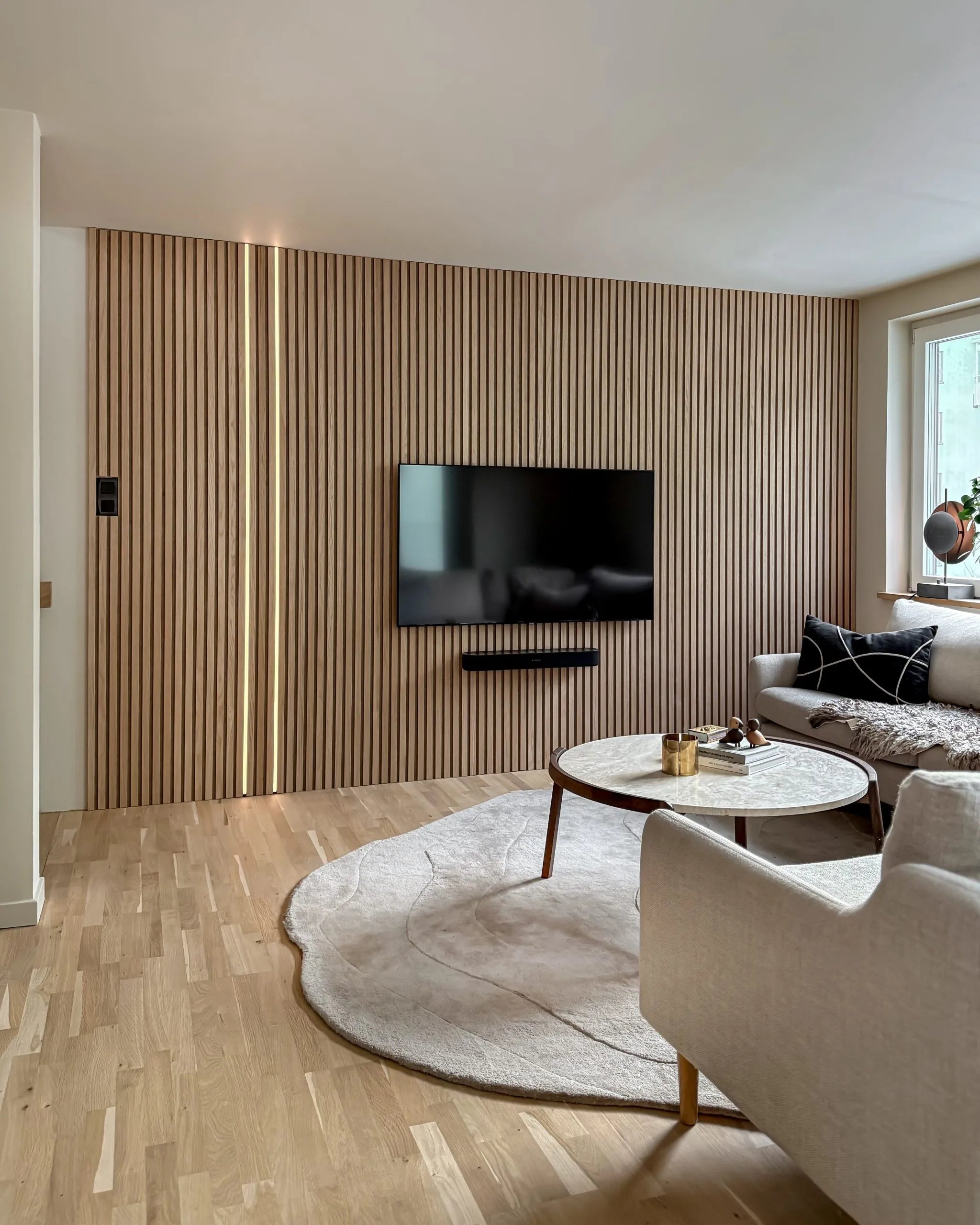 Moderne stue med en nordisk Akupanel akustikvæg af trælameller, monteret tv, rundt sofabord, cremefarvet sofa, stort tæppe på lyst trægulv.