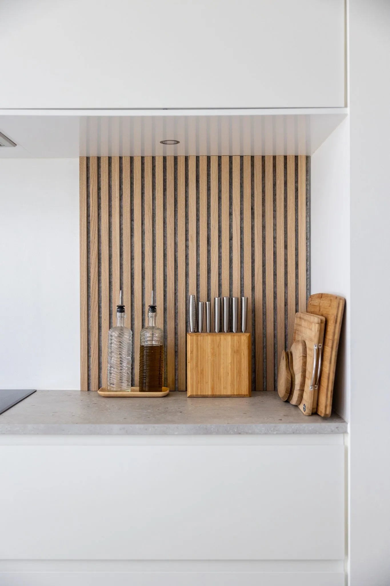 Moderne køkkenbord med olieflasker, knivblok i træ og stablede skærebrætter på baggrund af et nordisk Akupanel-backsplash med akustiske trælameller og hvide skabe.