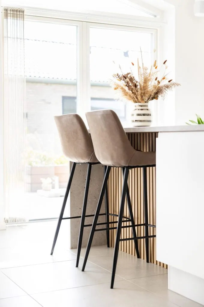 Beige barstole ved en køkkenø med en vase med tørrede blomster, store vinduer og nordisk moderne stil med akustiske paneler på væggene.