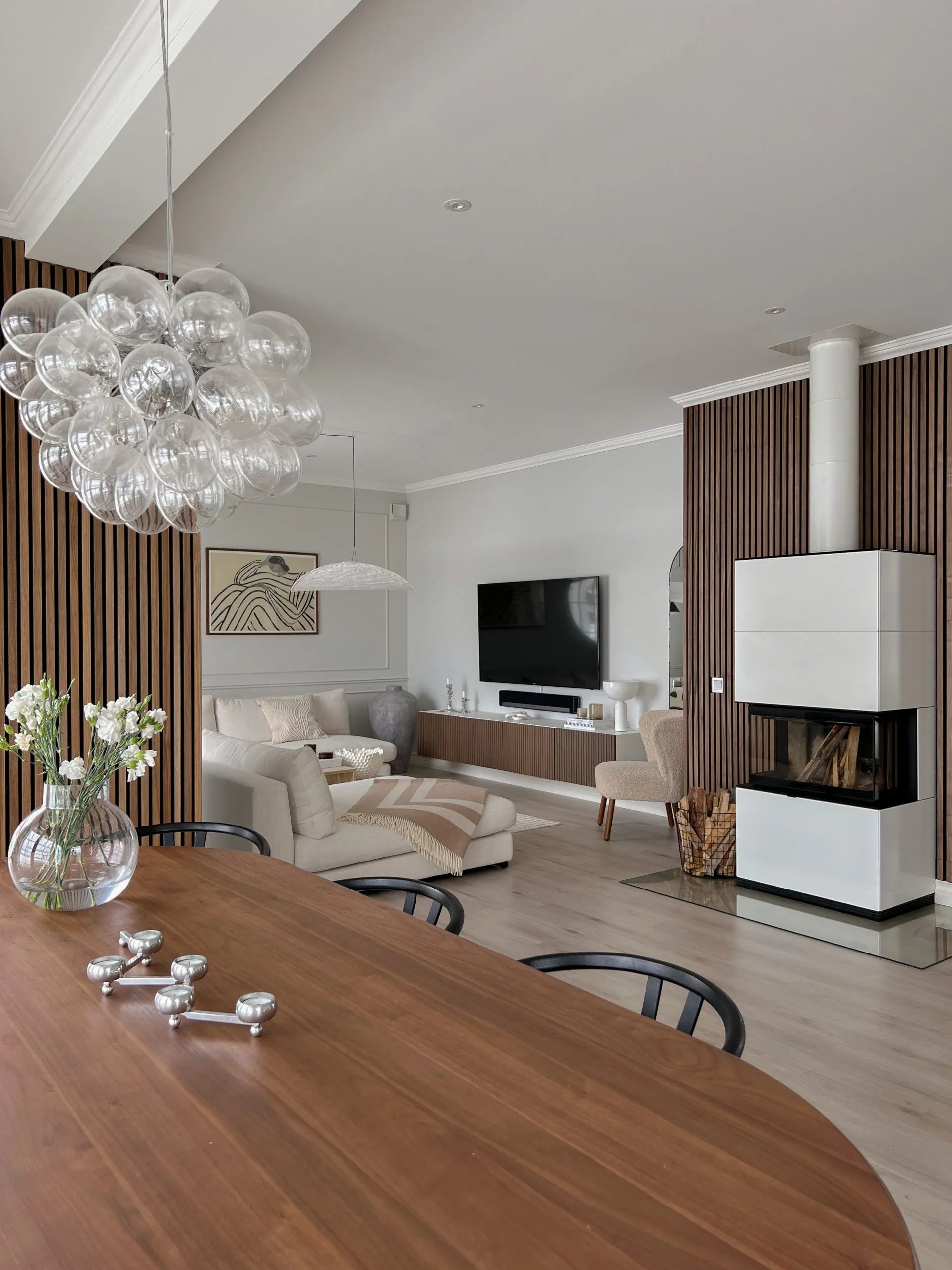 Moderne opholds- og spiseområde med akustiske paneler af nordisk træ, hyggelige siddepladser, tv, elegant pejs med stablet brænde og en lysekrone med glasbobler.