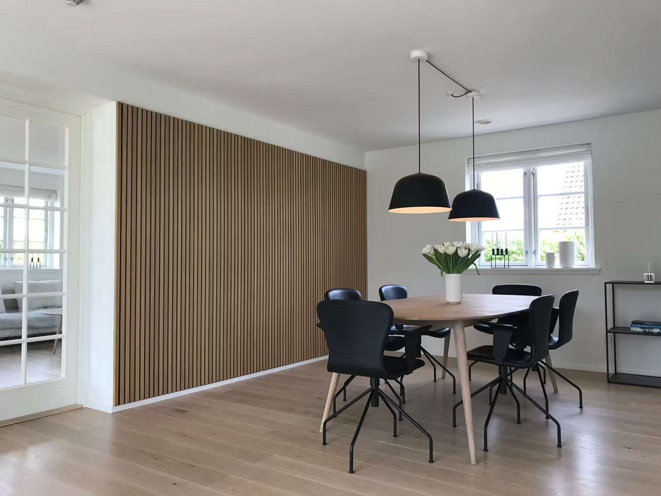 Moderne spisestue med trægulv, Akupanel akustisk lamelvæg, sorte stole, træbord, pendellamper og en hvid blomsterdekoration.