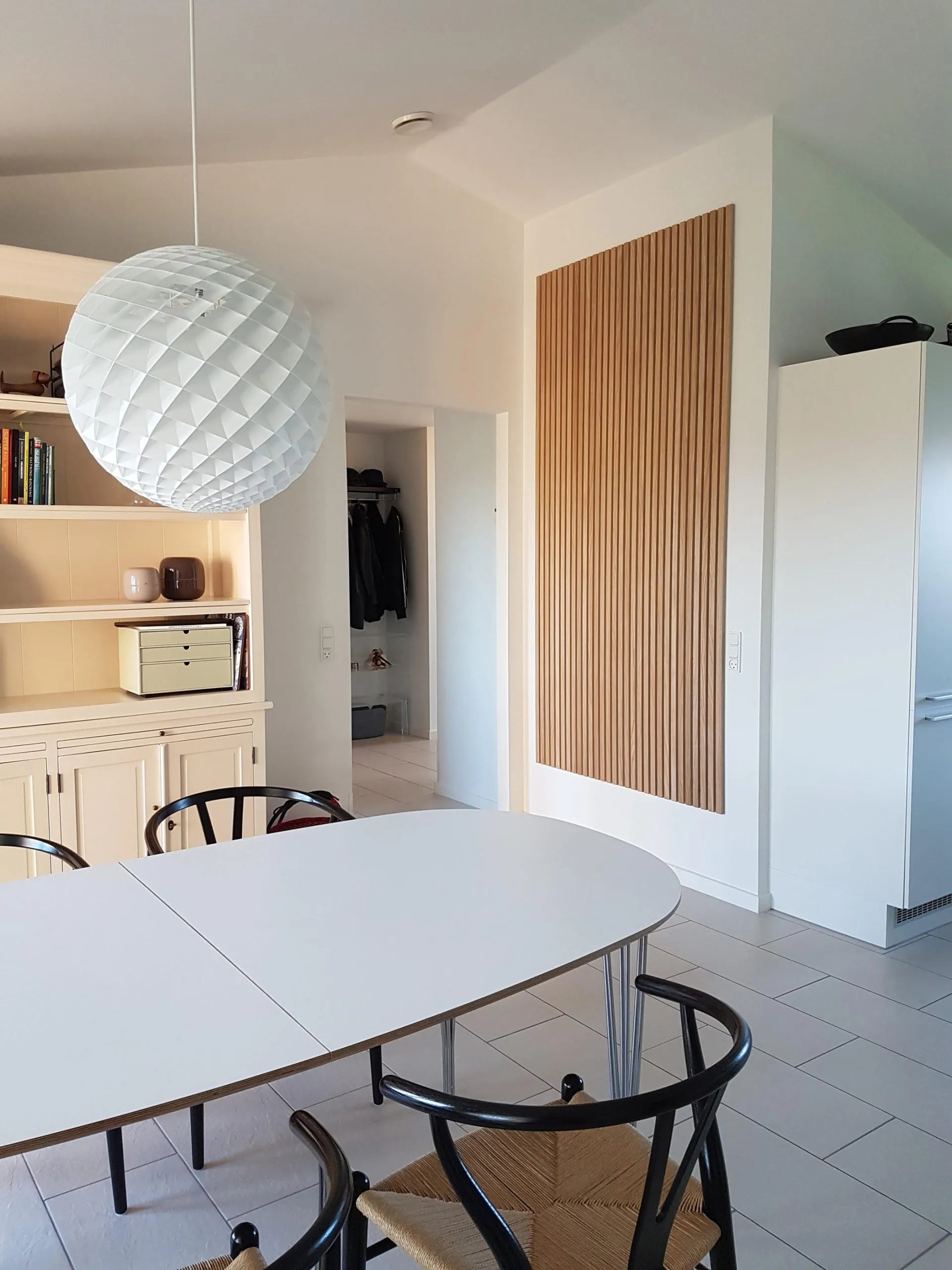 Moderne spiseplads med et hvidt ovalt bord, sorte stole, en stor struktureret pendel og slanke akustiske Akupanel-skabe i lyst træ.