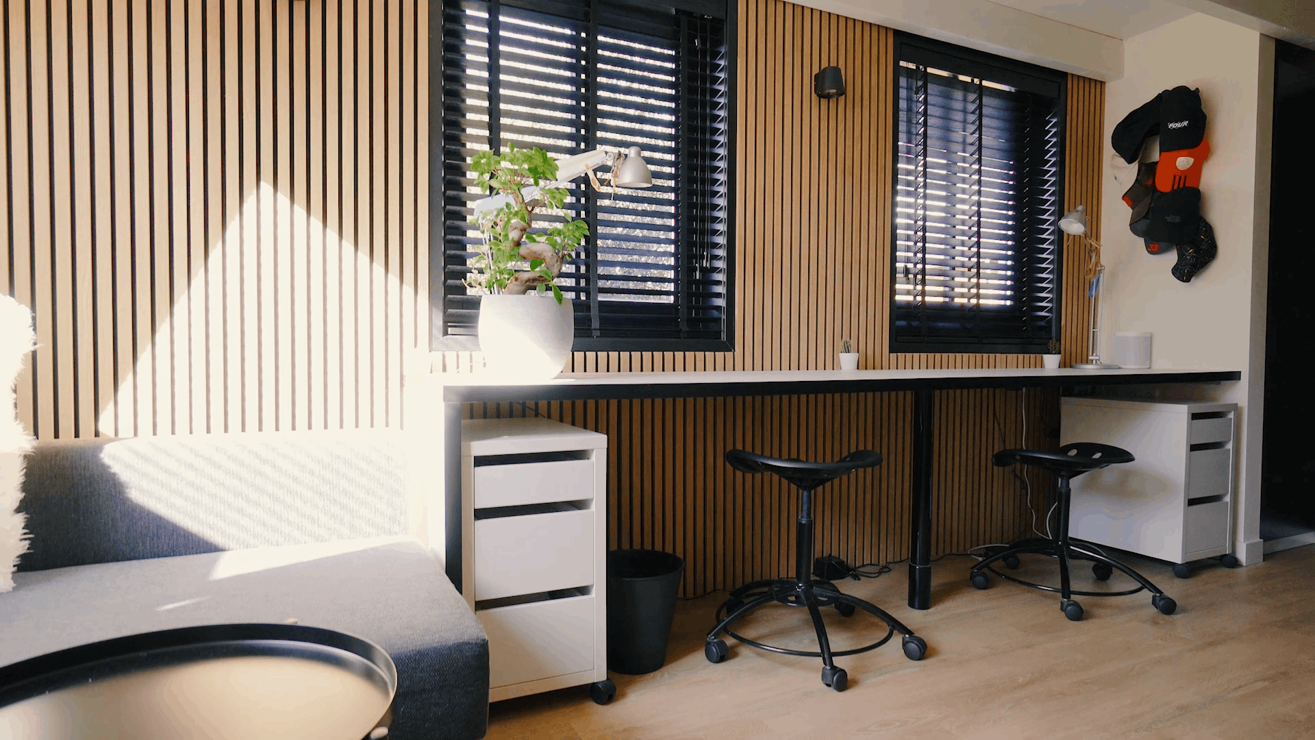 Moderne kontor med nordiske akustiske træpaneler på væggene, ergonomiske arbejdsstationer, sorte taburetter, en potteplante og sollys gennem sorte persienner.
