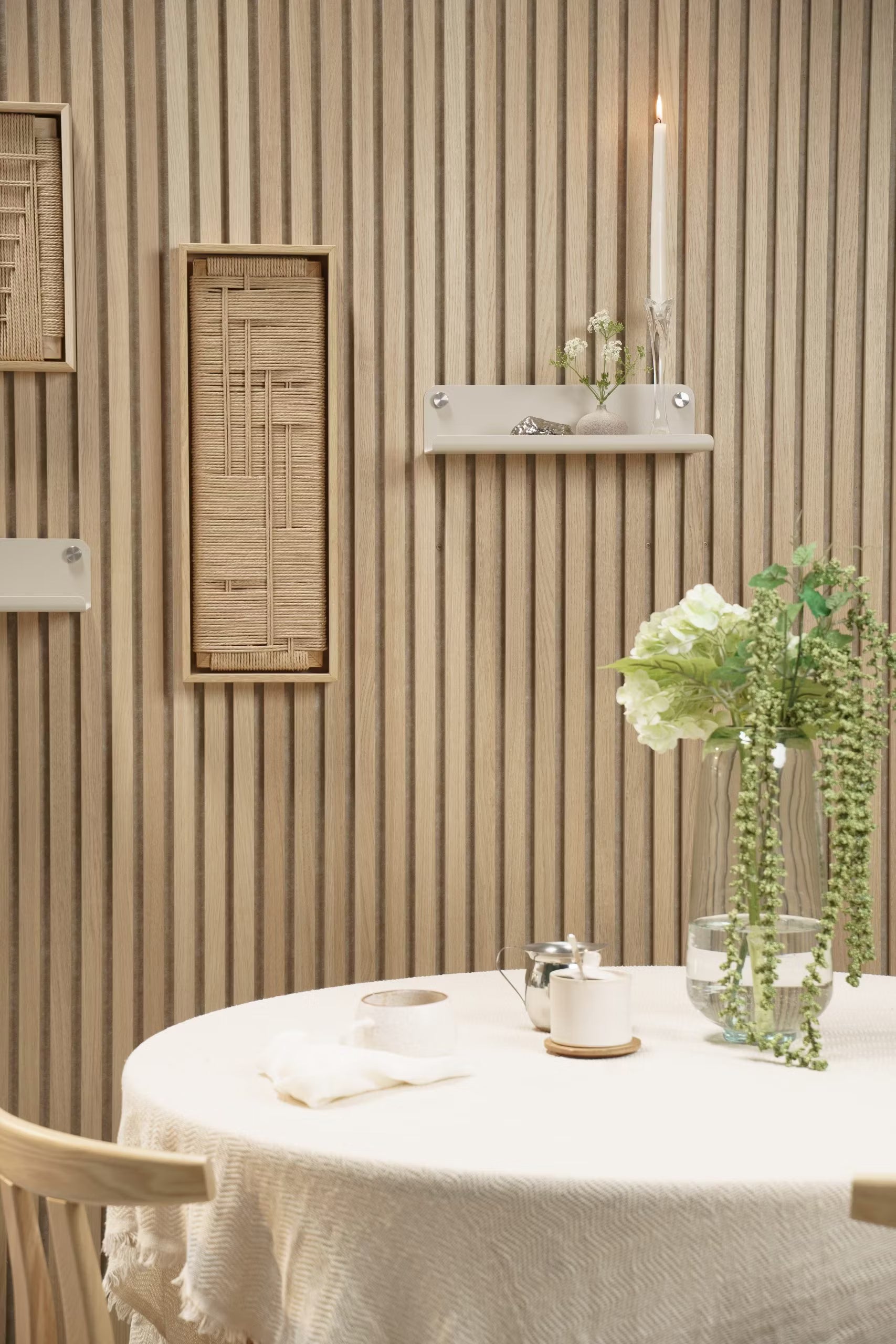 Et rundt bord med en hvid dug, blomstervase, stearinlys og dekoration placeret foran en Akupanel akustikvæg i lyst træ med vævede og hyldeagtige accenter.