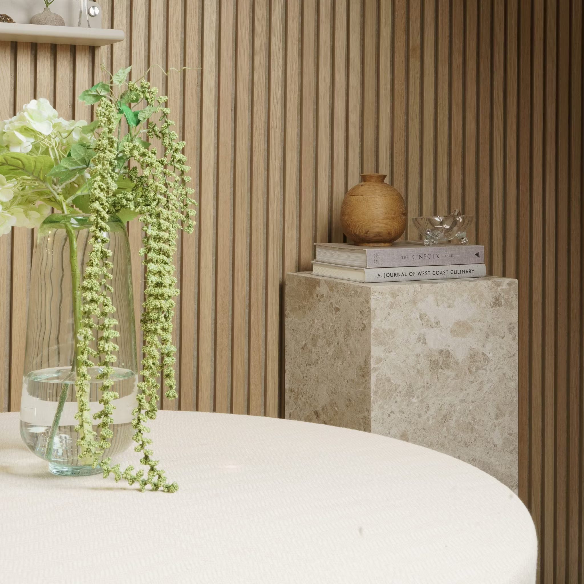 En vase med blomster står på et rundt bord; bøger og keramik hviler på en marmorsokkel mod en Akupanel akustisk træpanelvæg.