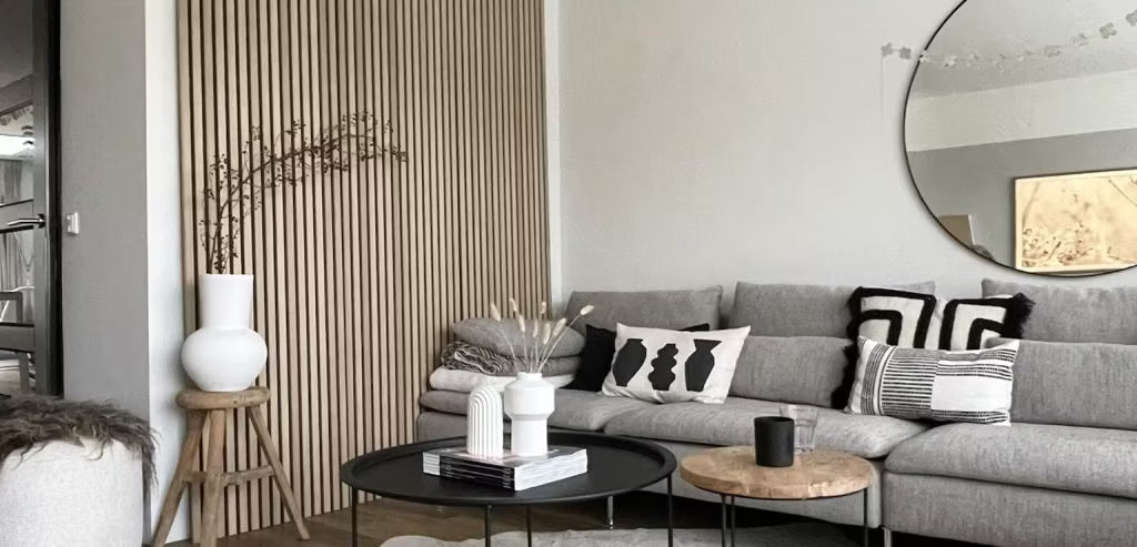 Moderne stue med en grå sofa, abstrakte puder, sorte sofaborde, dekorative vaser og en akustisk Akupanel-væg af træ med et rundt spejl.