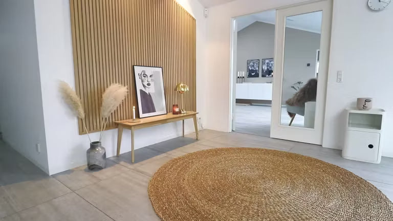 Minimalt nordisk opholdsrum med et rundt jutetæppe, en træbænk med pampasgræs, indrammet kunst og en Akupanel akustisk trævæg med lameller.