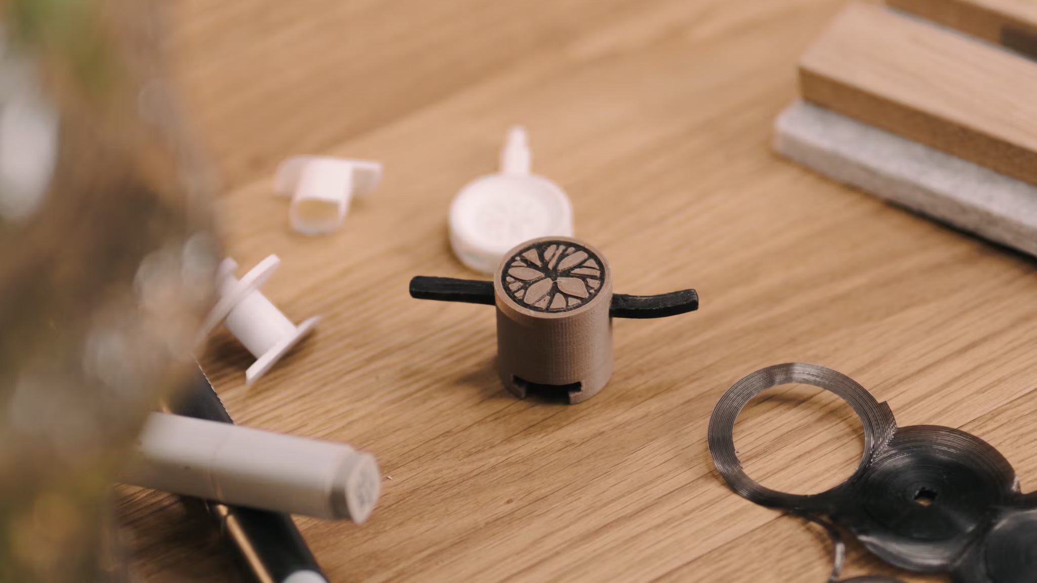 En lille 3D-printet prototype med sorte arme ligger på et træbord ved siden af forskellige plastikkomponenter, der refererer til udviklingen af tilbehør til akustikpaneler.