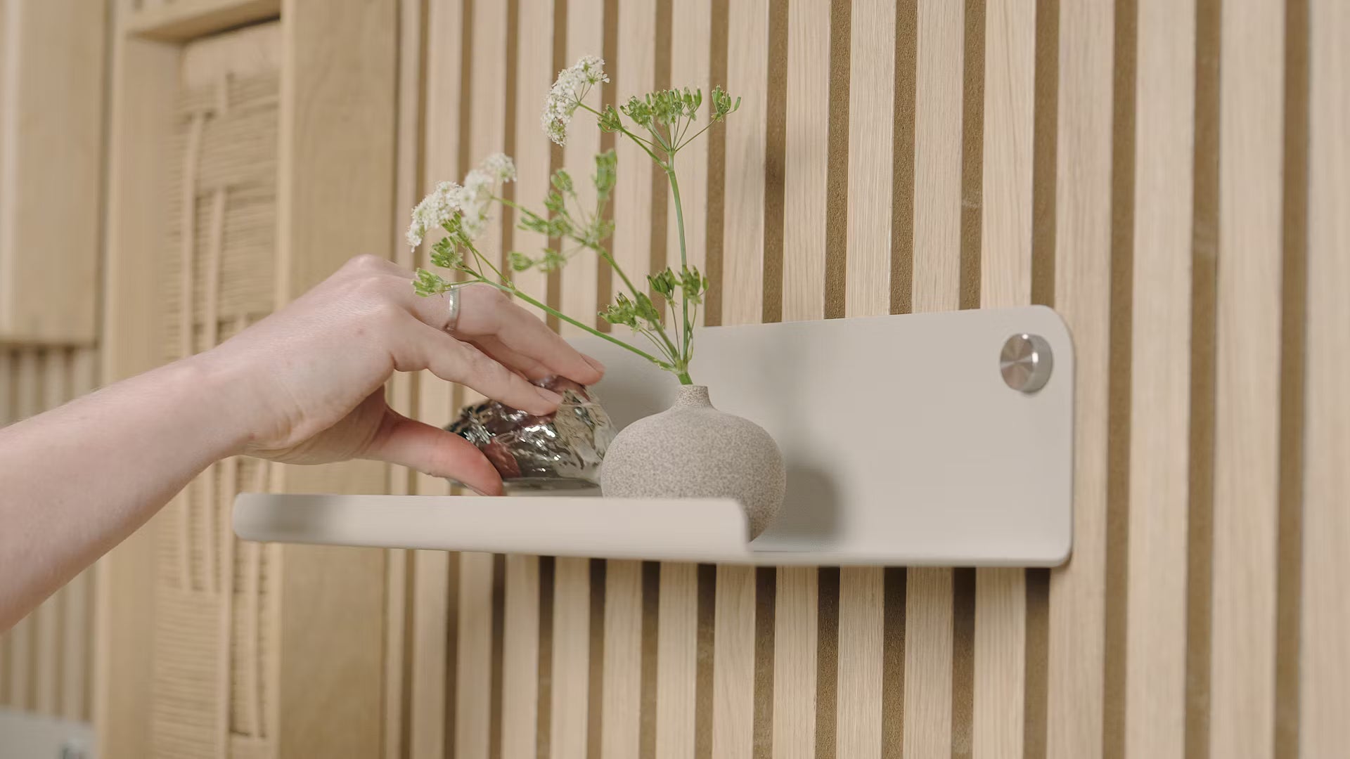 En hånd placerer et lille dekorativt objekt ved siden af en vase med hvide blomster på en minimalistisk hylde, der står op ad et struktureret nordisk akustisk træpanel.