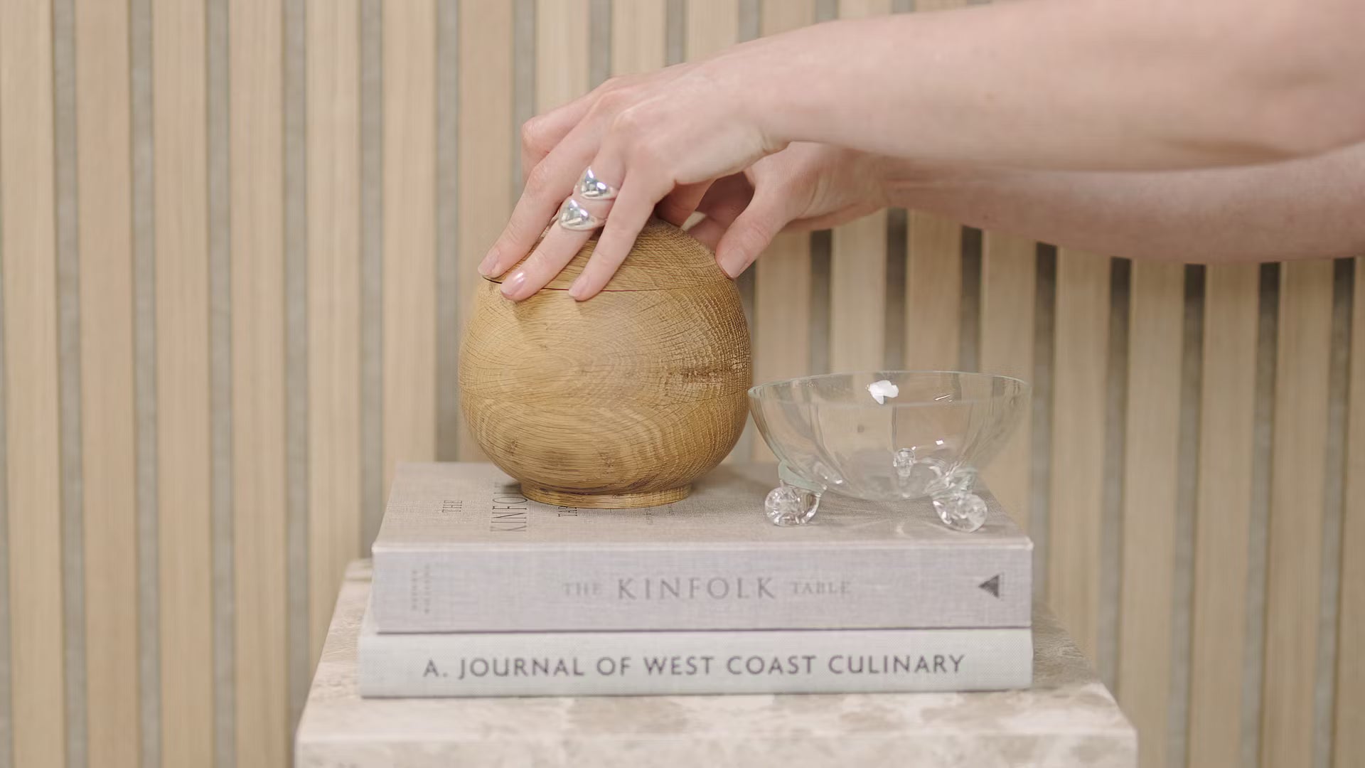 En hånd rører ved en trækugle, der er placeret på stablede bøger, herunder Kinfolk Table og Journal of West Coast Culinary, ved siden af en glasskål på et akustisk panel af trælameller.