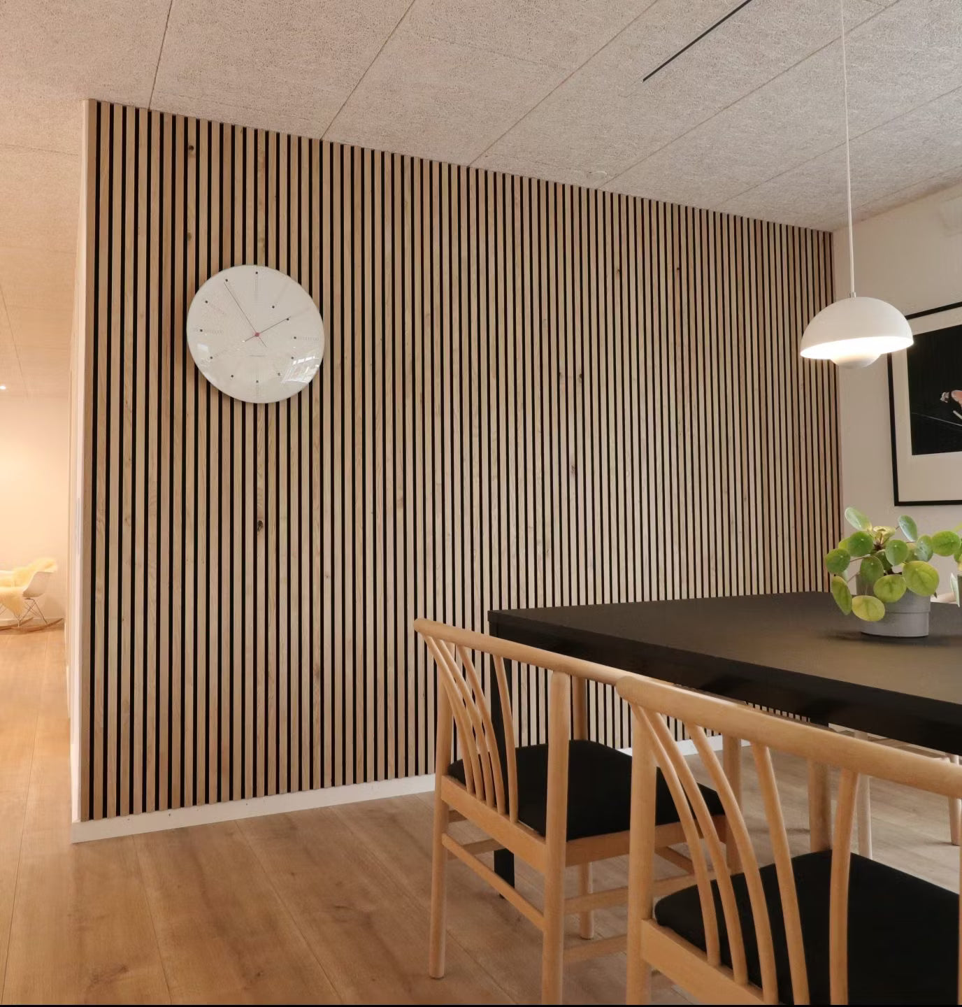 Moderne spisestue med en nordisk Akupanel akustisk træpladevæg, sort bord, træstole med sorte sæder og en potteplante i midten.
