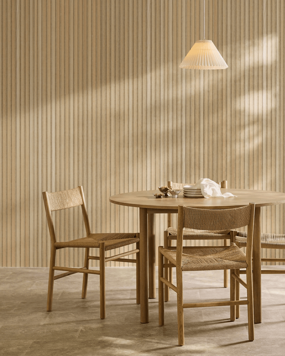 Minimalistisk spiseområde med et rundt bord, flettede stole og en pendellampe ved siden af et WoodUpp Akupanel akustikpanel i træ i Classic Oak med beige filt.