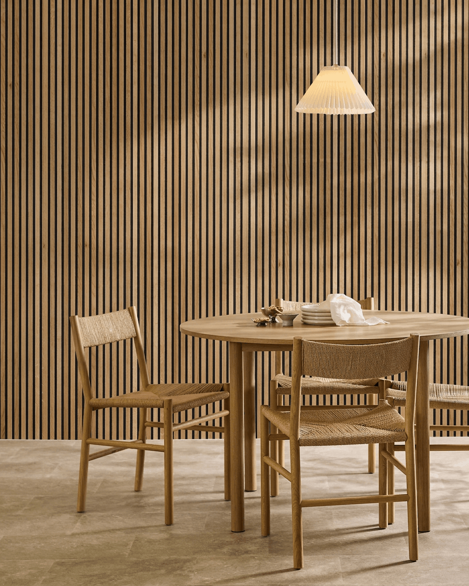 Minimalistisk spiseområde med et træbord, to stole og et WoodUpp Akupanel akustisk træpanel i Rustic Brown Oak (300x60 cm), oplyst af en hængelampe.