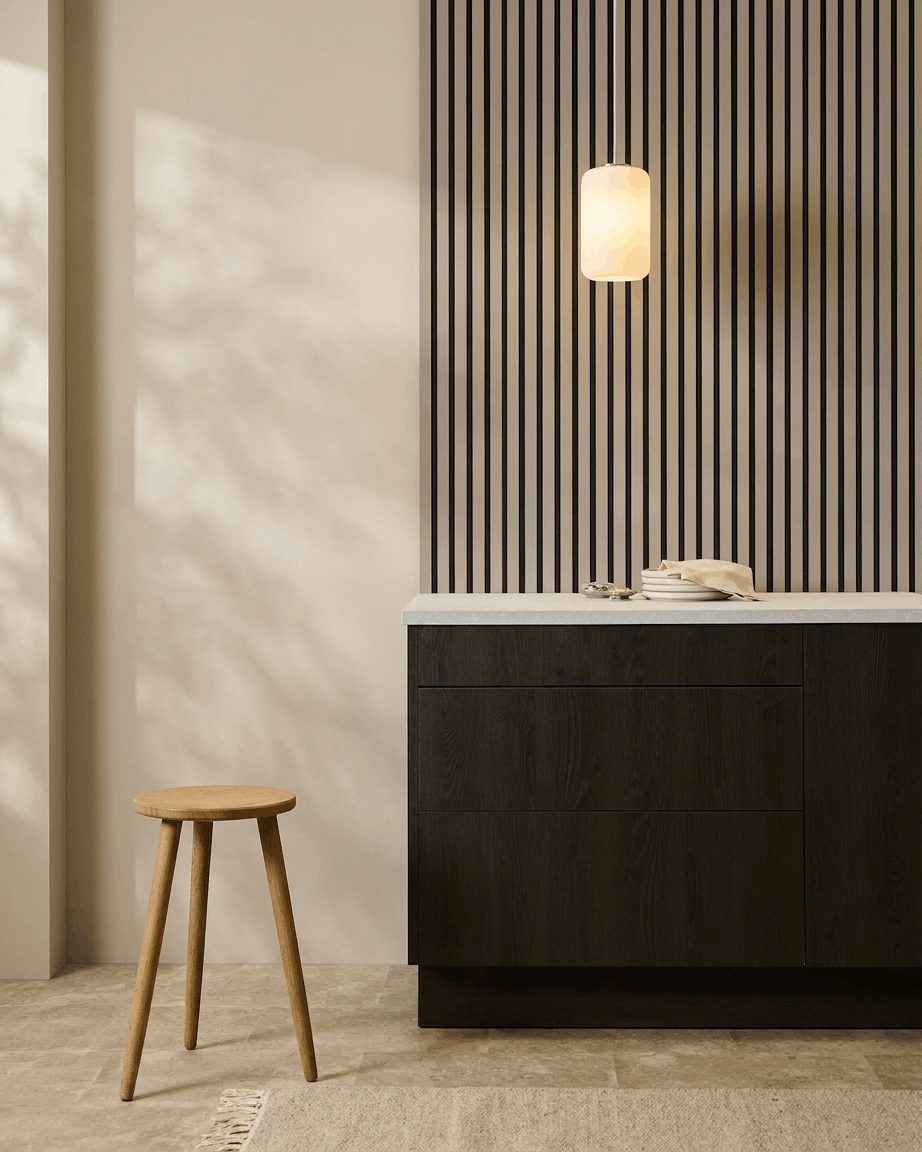 Minimalistisk køkken med WoodUpp Akupanel (Stone, Black filt) akustikpaneler på væggen, træskammel, mørke skabe og pendellampe i neutrale toner.