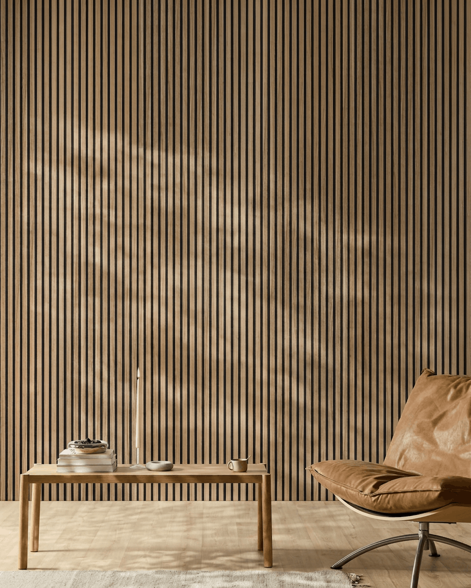 Minimalistisk rum med et WoodUpp Akupanel Akustikpanel (English Walnut-lameller, black filt), en stol i tanfarvet læder, et bord i lyst træ, et stearinlys, bøger, en kop og et hvidt tæppe i sollys.