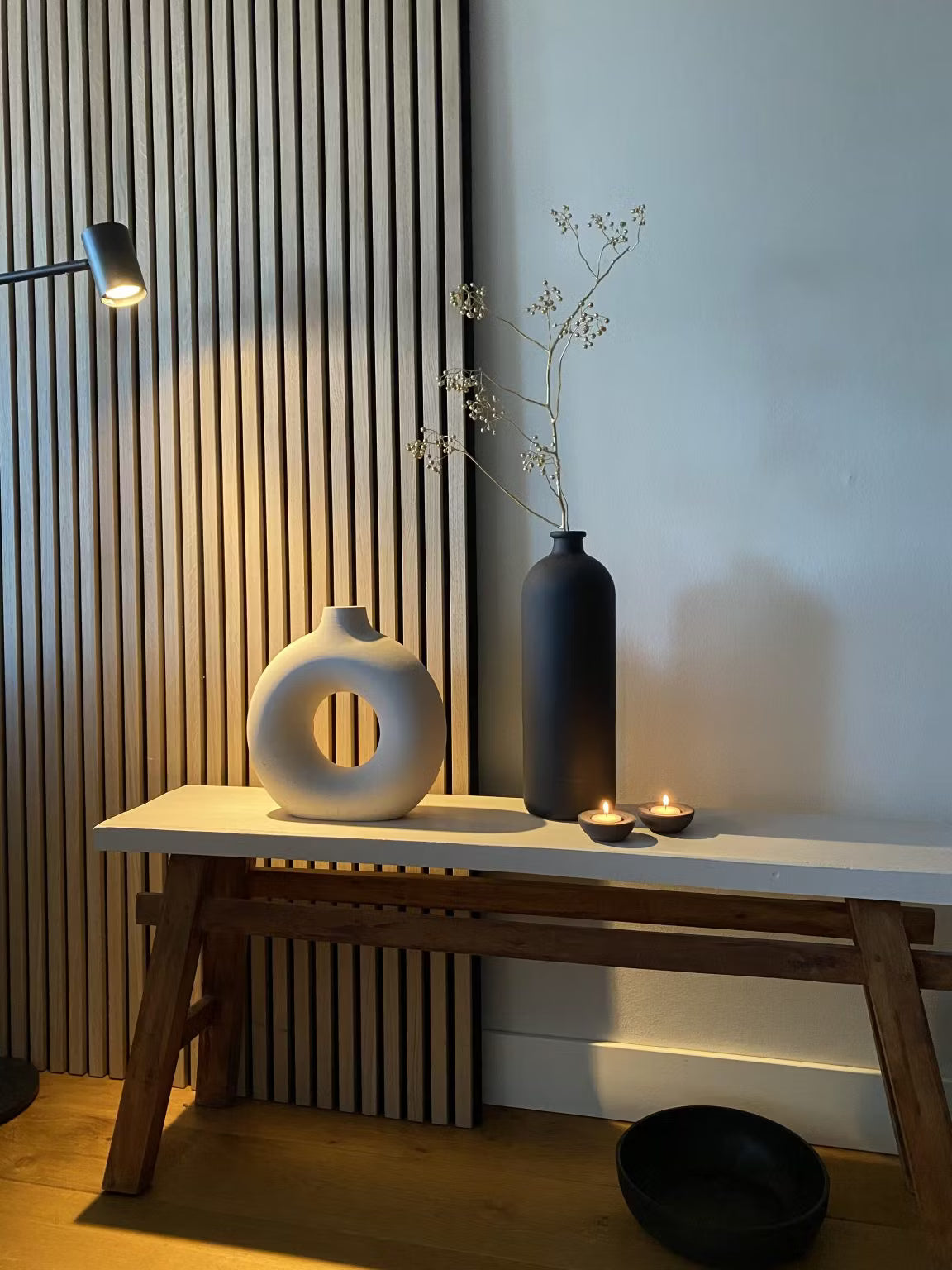 Minimalistisk indretning med en hvid cirkulær vase, en sort høj vase med tørrede stilke, to fyrfadslys og en sort skål på baggrund af Akupanel-træplader.
