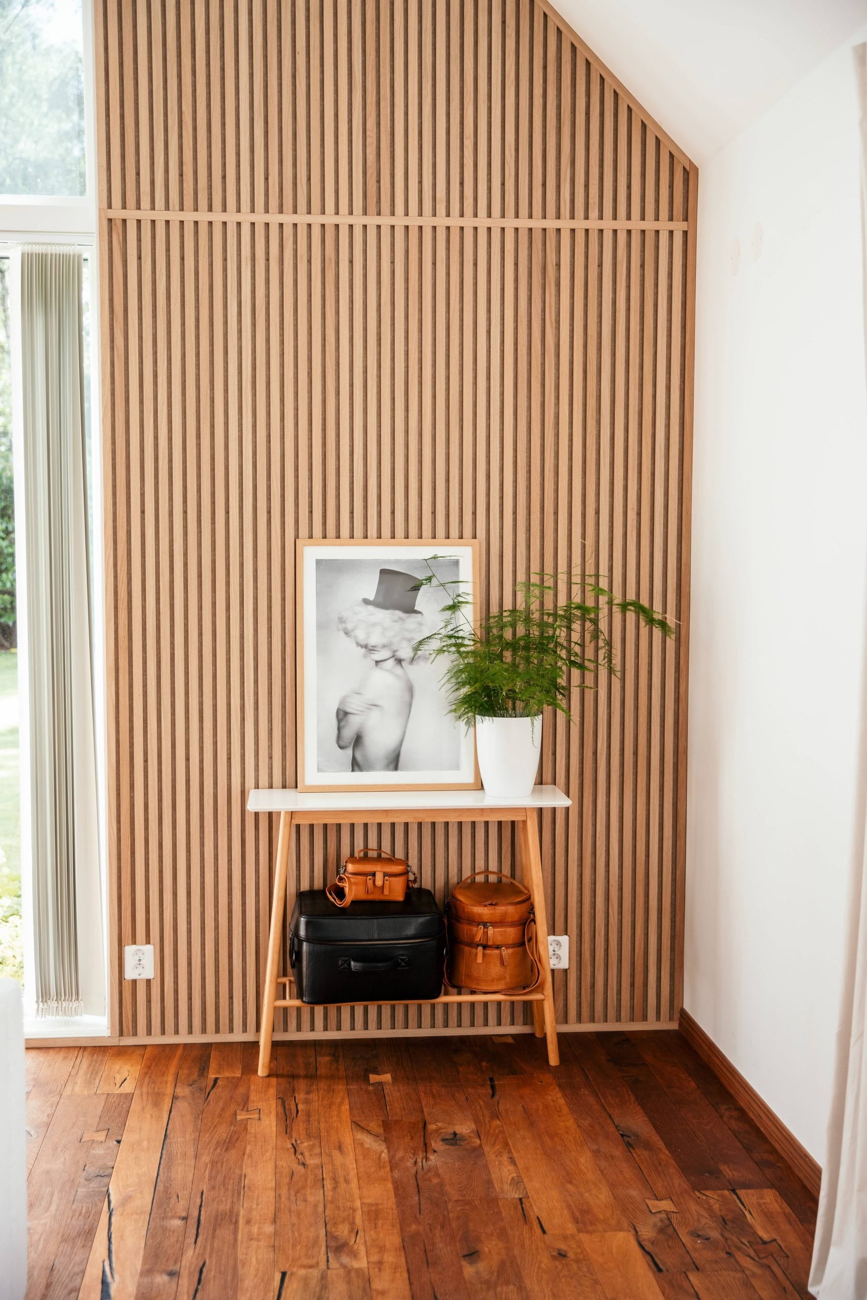 Et lille træbord med tasker, et indrammet foto og en potteplante står op ad en nordisk akustisk lamelvæg i et lyst rum med trægulv.