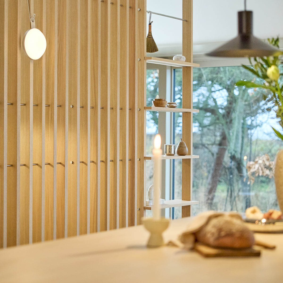 Minimalistisk interiør med en Nordic EMBRACE trærumdeler, tændt lys, frisk brød på bordet, udvalgt pynt på hylderne og et vindue, der indrammer træer udenfor.