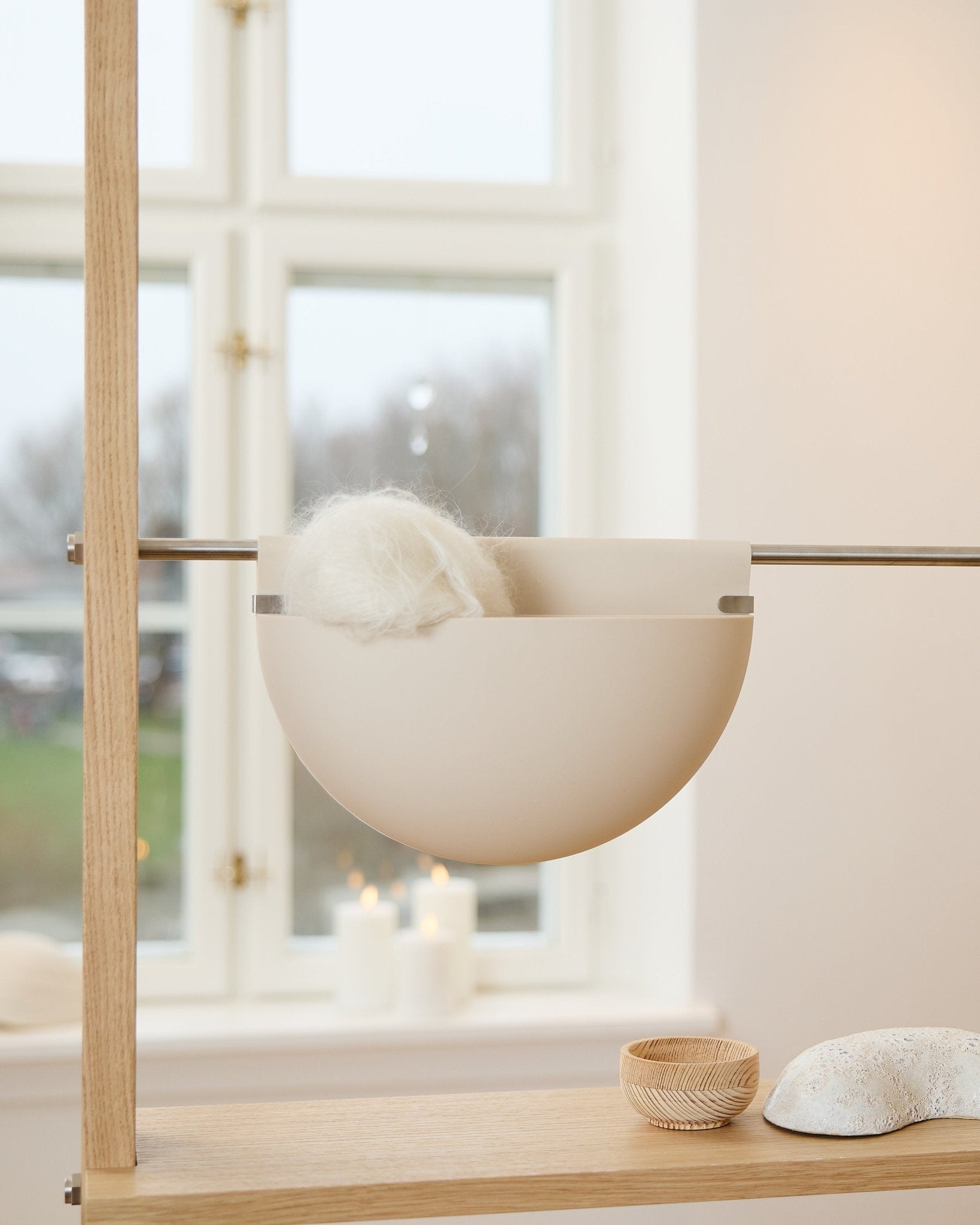 A white bowl with a fluffy object rests on a wooden shelf near candles, decor, and a WoodUpp Gro Soft Pocket BM2 acoustic accessory in light beige by the window.