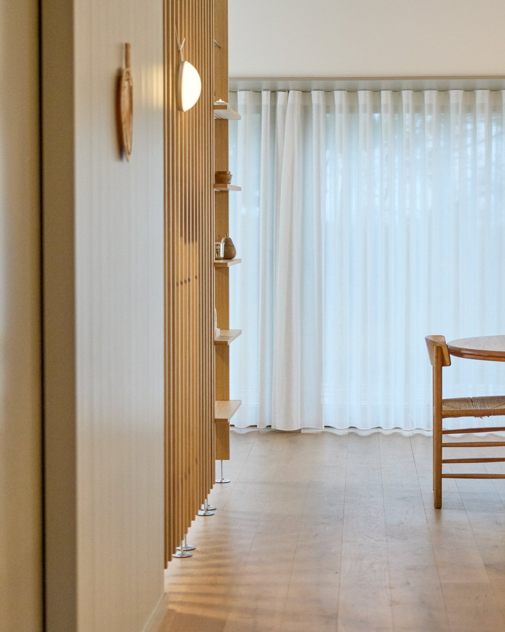 Minimalistisk rum med en WoodUpp Embrace rumdeler fra gulv til loft, trægulv, hylder, stol og transparente gardiner foran large vinduer.