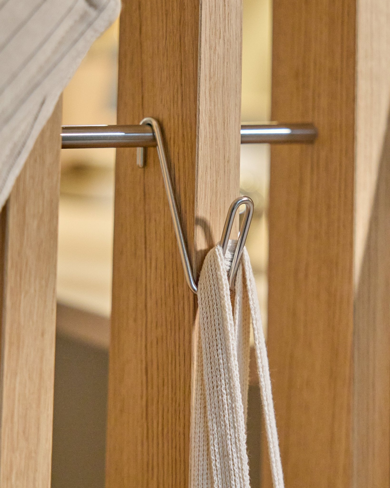 The Alva Wire Hook / BM1 (Stainless Steel, Short) by WoodUpp is mounted on a wooden acoustic panel rail, holding a white strap with metal rod; beige backdrop.