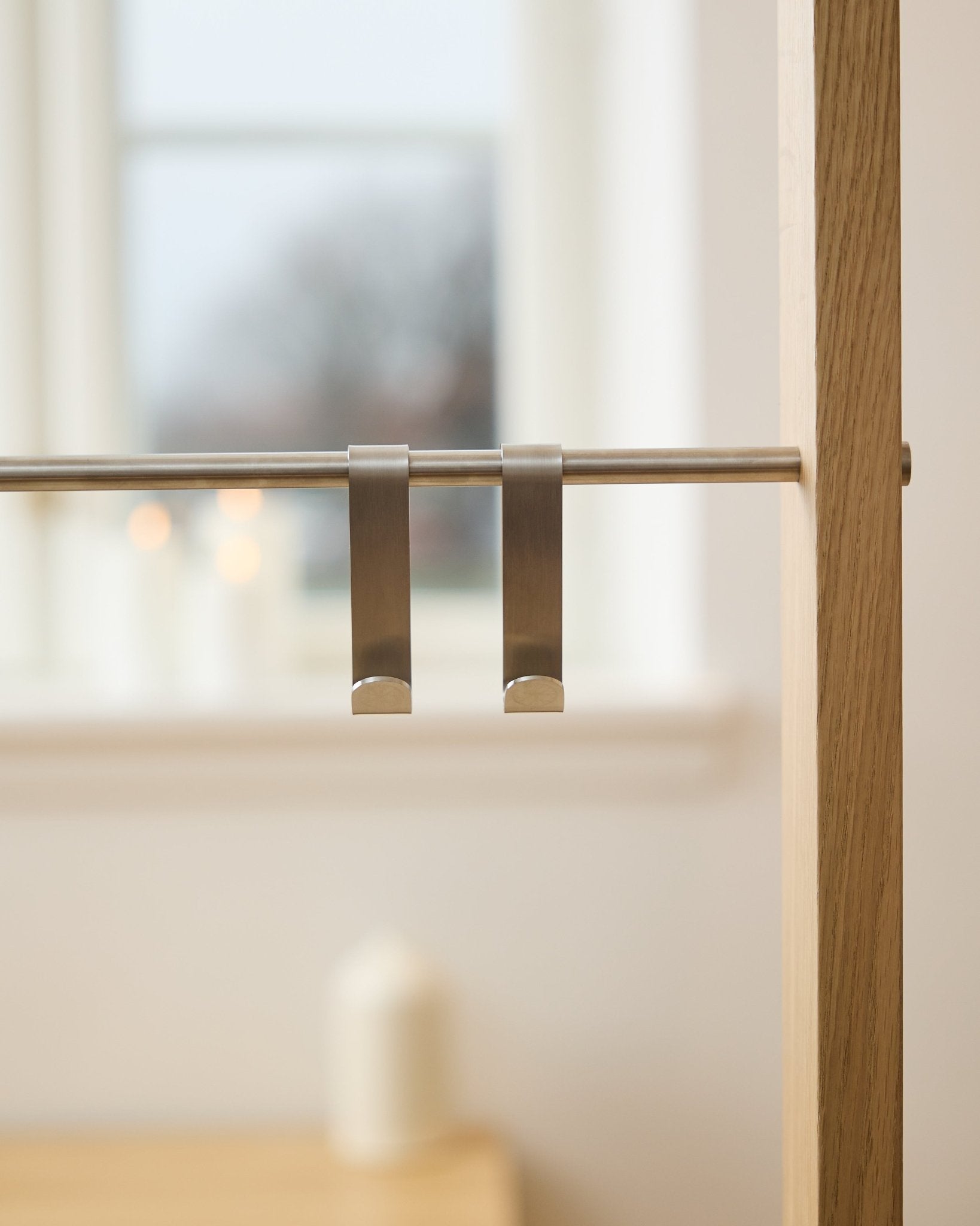 WoodUpp Alva S-krog BM1  BM2 (2 stk.) i rustfrit steel, hænger på en stang til akustikpanel i træ; sløret vindue og tændte stearinlys i baggrunden.