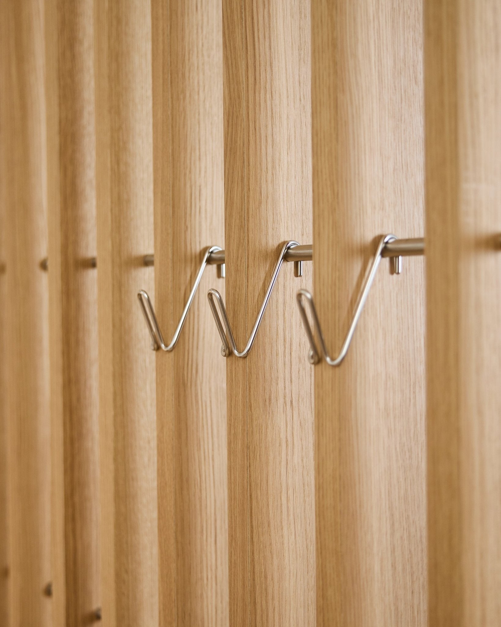 Tre korte Alva Wire Hook / BM1 accessories fra WoodUpp i rustfrit steel er monteret med jævn afstand på række på et vertical akustikpanel med trælameller.