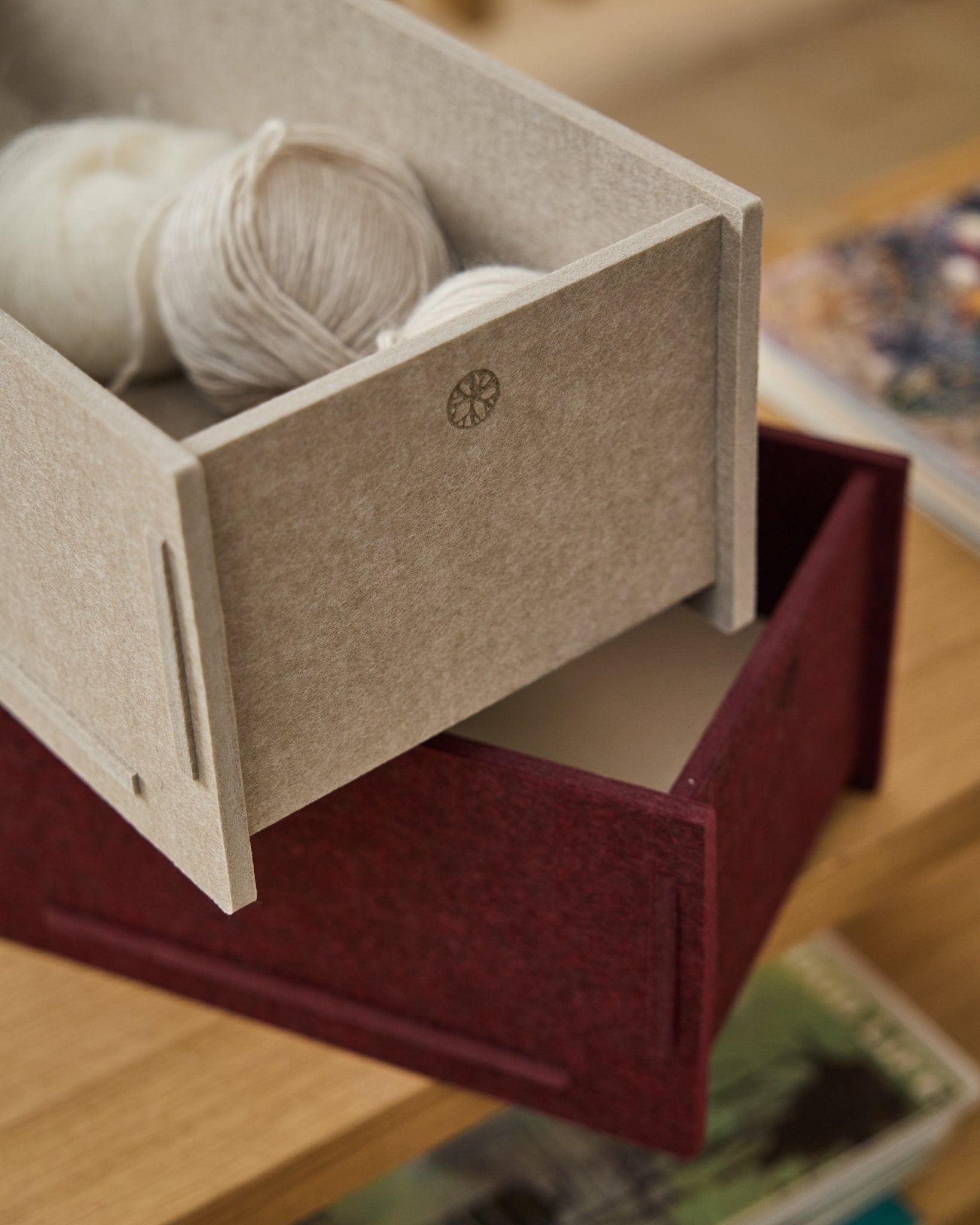 En beige opbevaringskasse i PET-filt fyldt med garn står oven på en lukket WoodUpp Vigg Felt Box/BM2 i ox red på et trægulv med bøger i baggrunden.