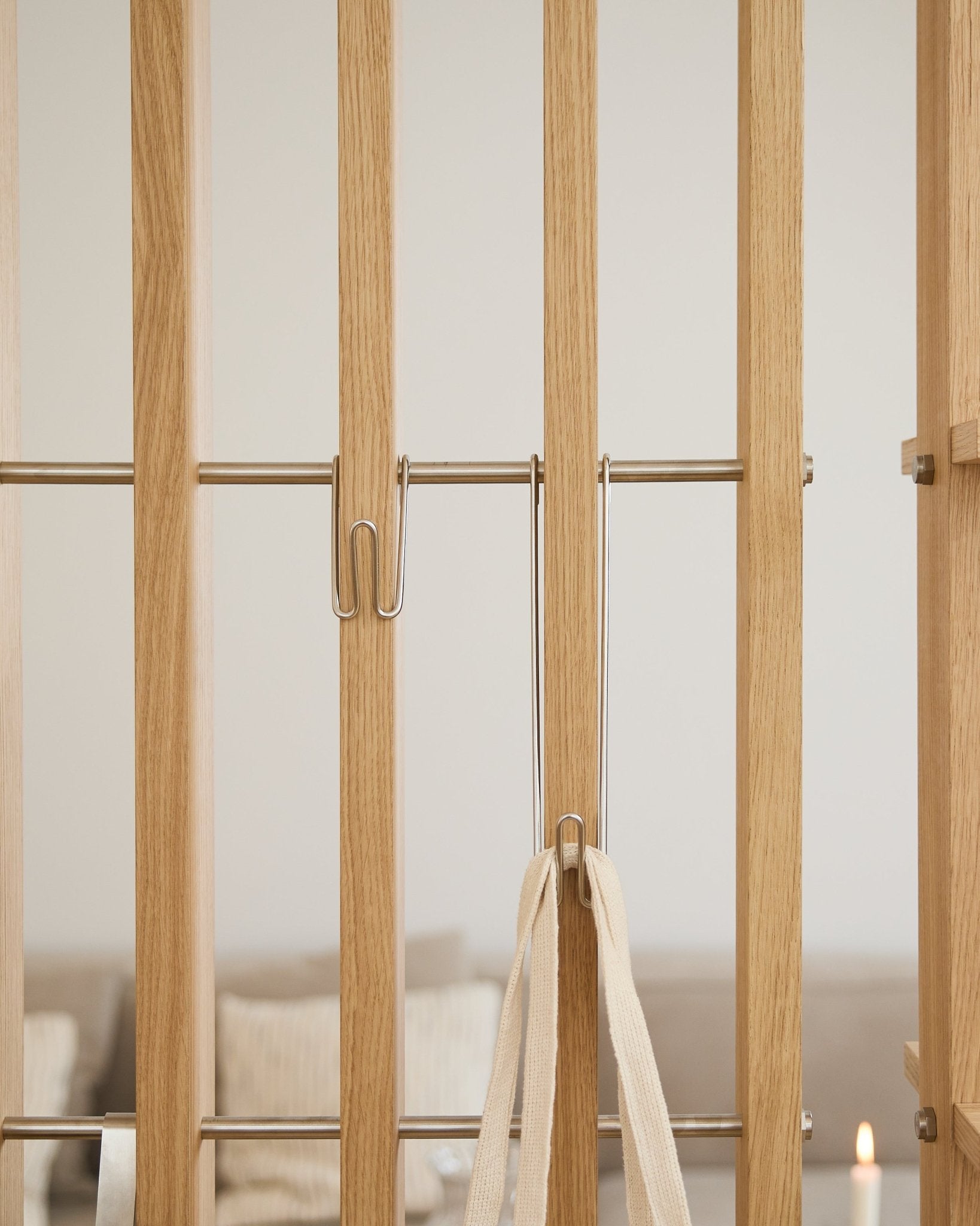 Close-up of WoodUpp Alva | Wire Hook BM1 (stainless steel, long, set of 3) attached to light acoustic panel slats with a white strap; sofa and candle blurred in background.