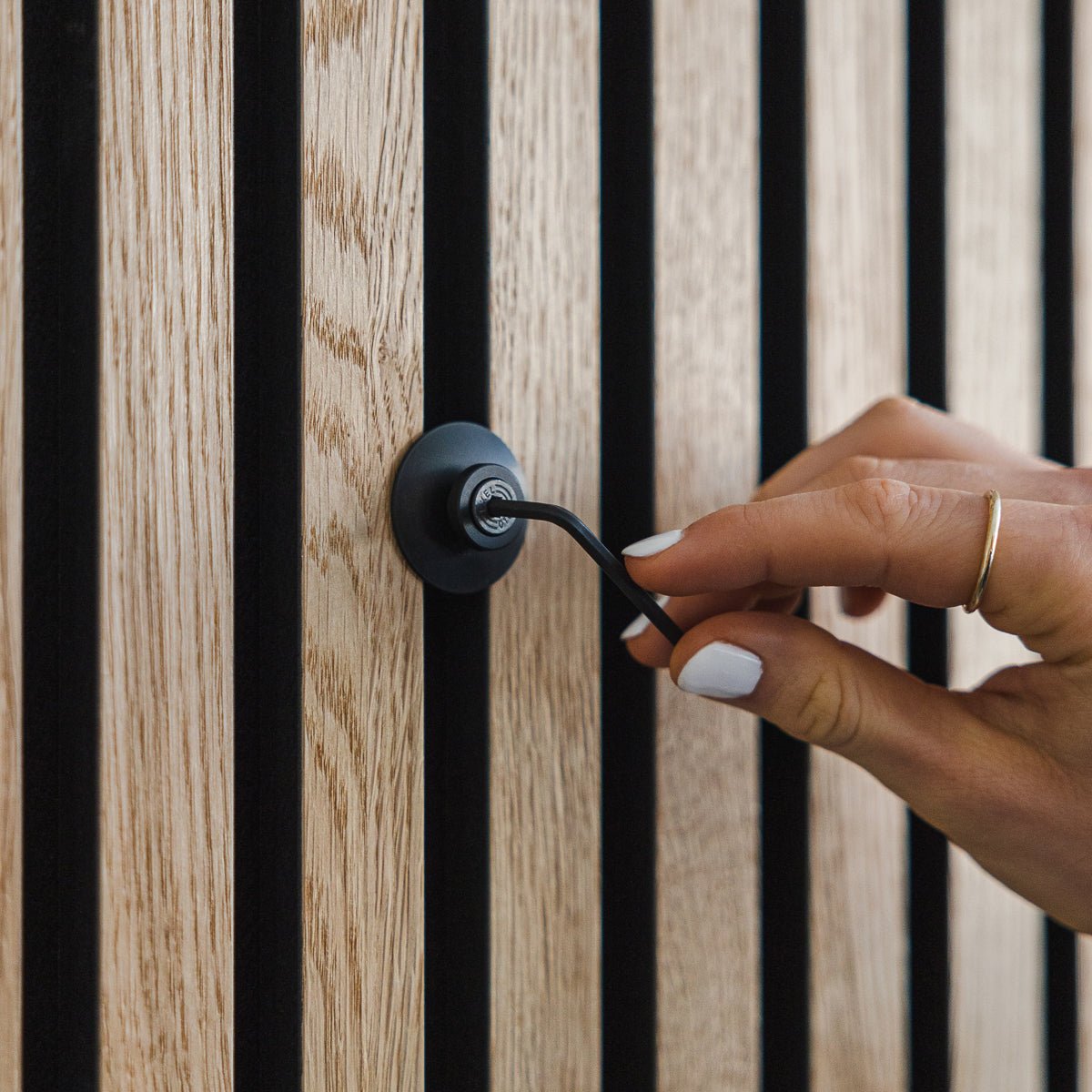 En hånd med hvid neglelak drejer en Dupps | Heavy black aluminiumskrog fra WoodUpp på et vertical Akupanel akustikpanel i træ.