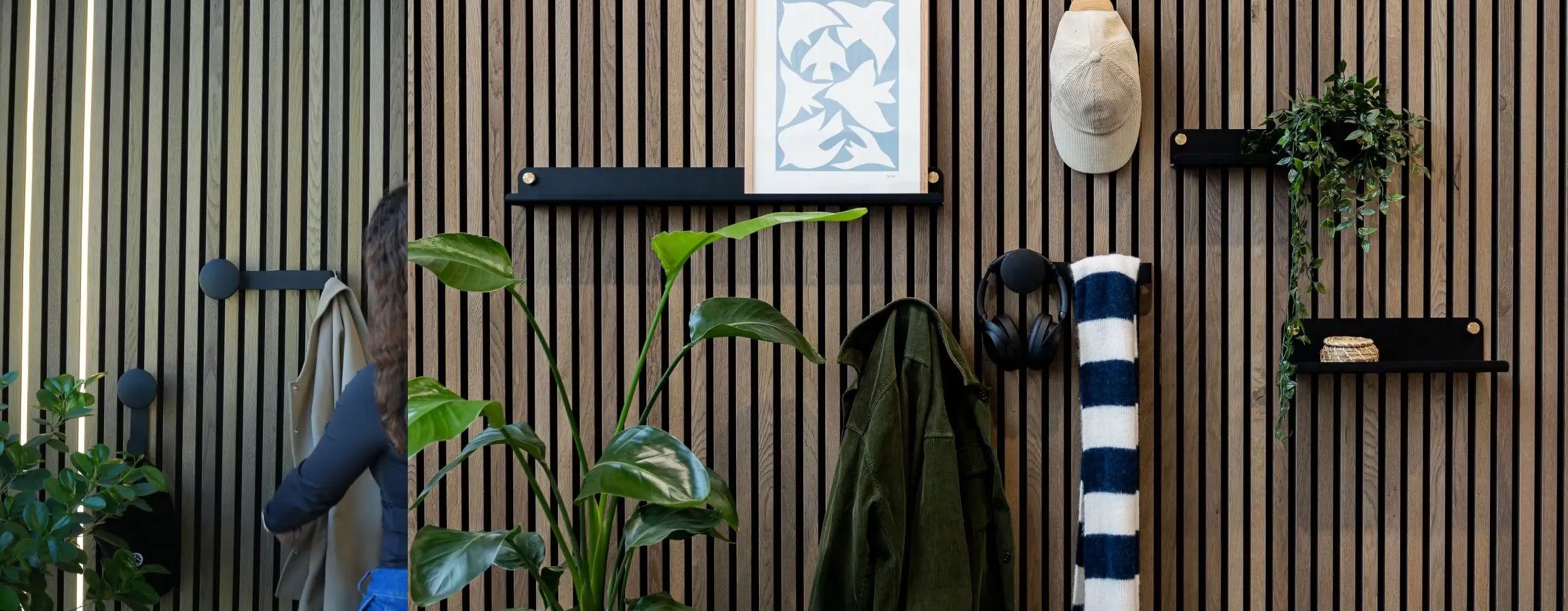 En mand i blå jakke går forbi en nordisk Akupanel akustisk træpladevæg med integrerede planter, hylder, knager, hovedtelefoner og et sammenrullet, stribet håndklæde.