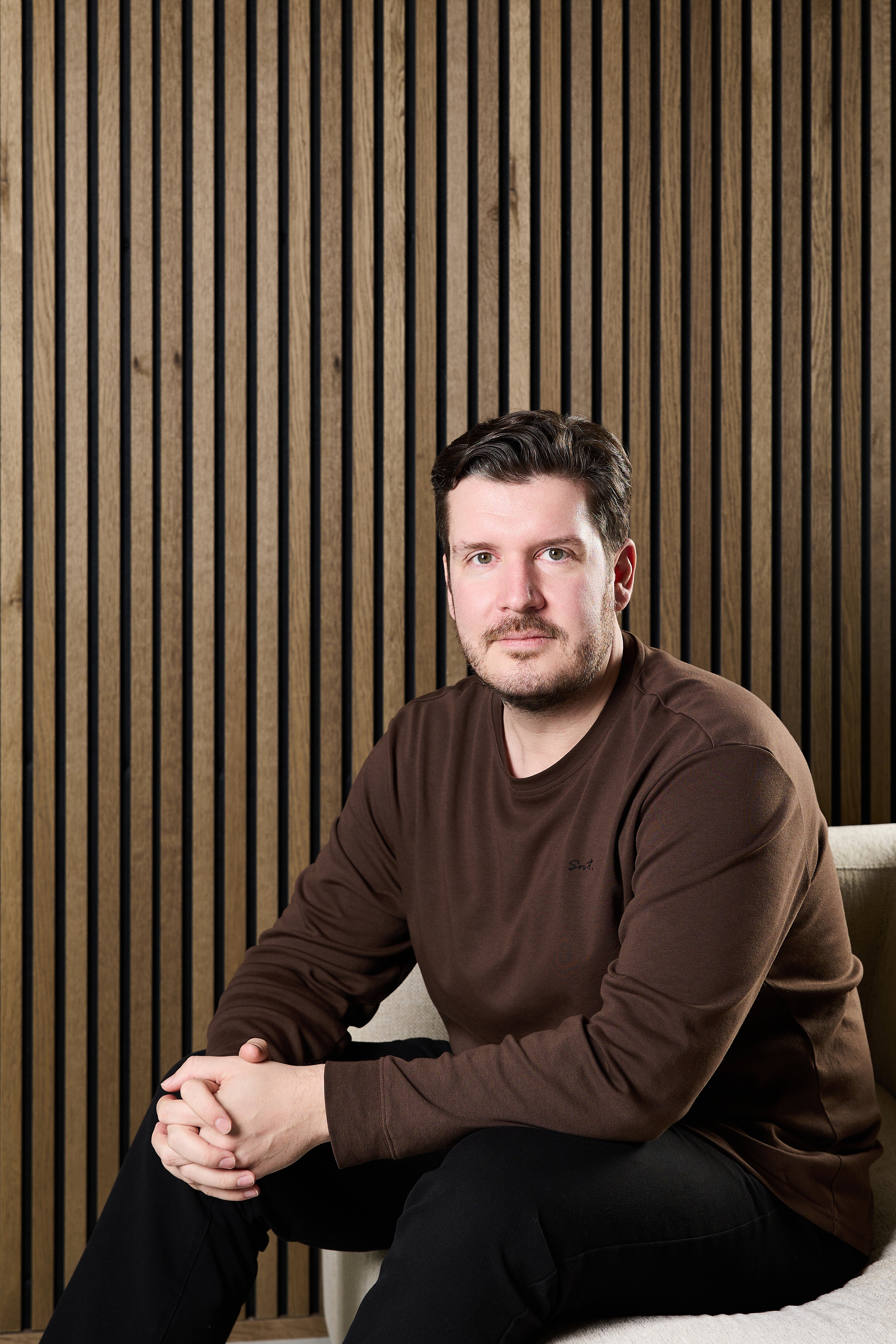 A man with short dark hair and a beard sits on a light chair in a brown sweater, positioned in front of a Nordic-style acoustic slatwall panel.