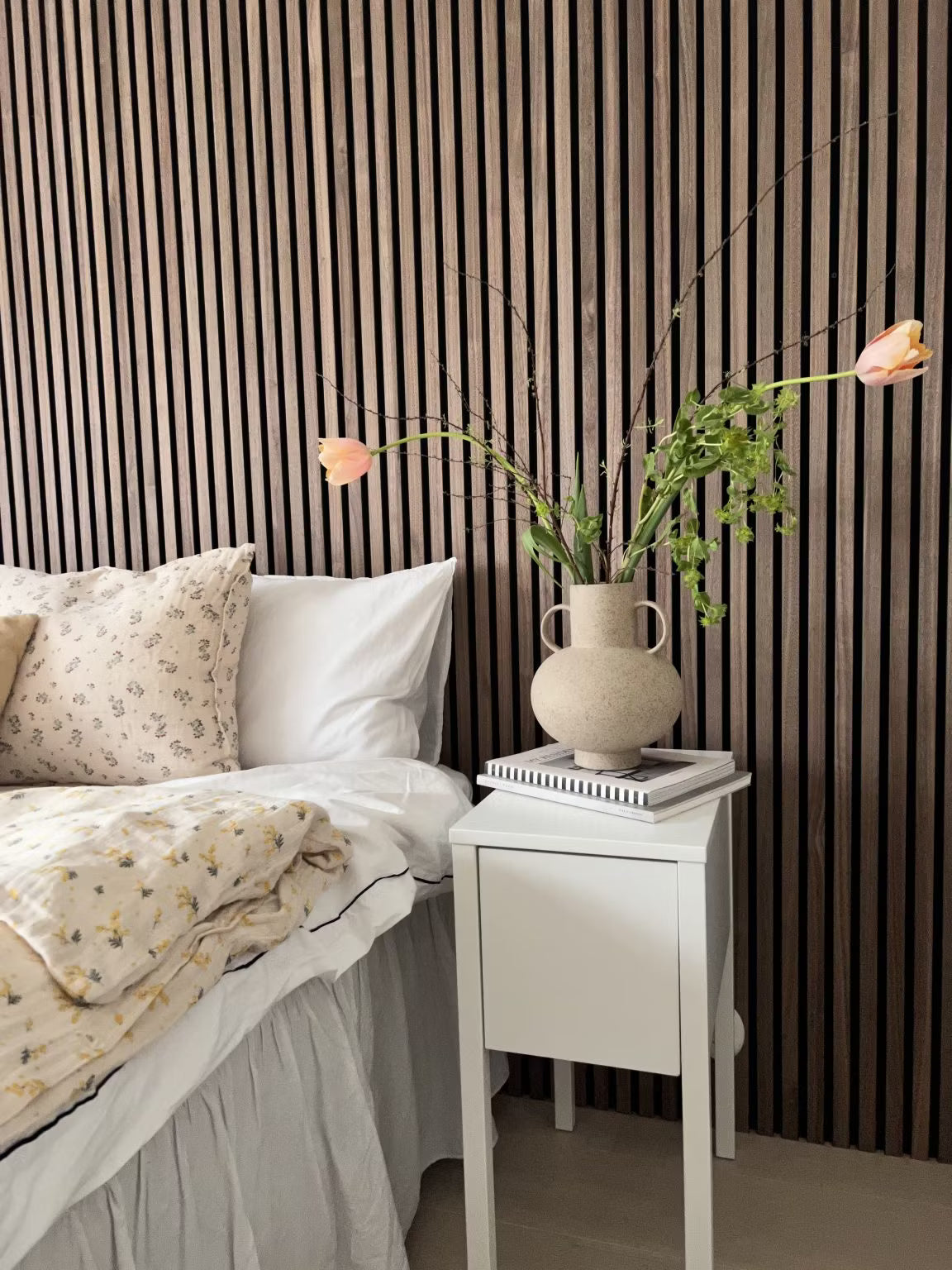 Et soveværelse med en hvid seng med blomstret sengetøj og en vase med blomster, med en nordisk Akupanel akustisk lamelvæg af træ bag sengen.