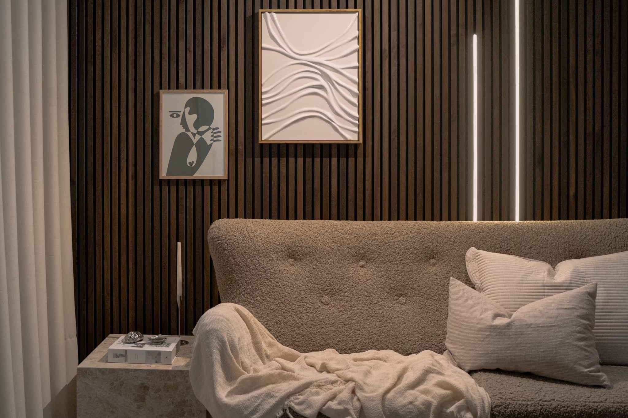 Moderne stue med struktureret beige sofa, hvide puder, abstrakt akustisk panelkunst og lodret Akupanel-trælistevæg med blød, stemningsfuld belysning.
