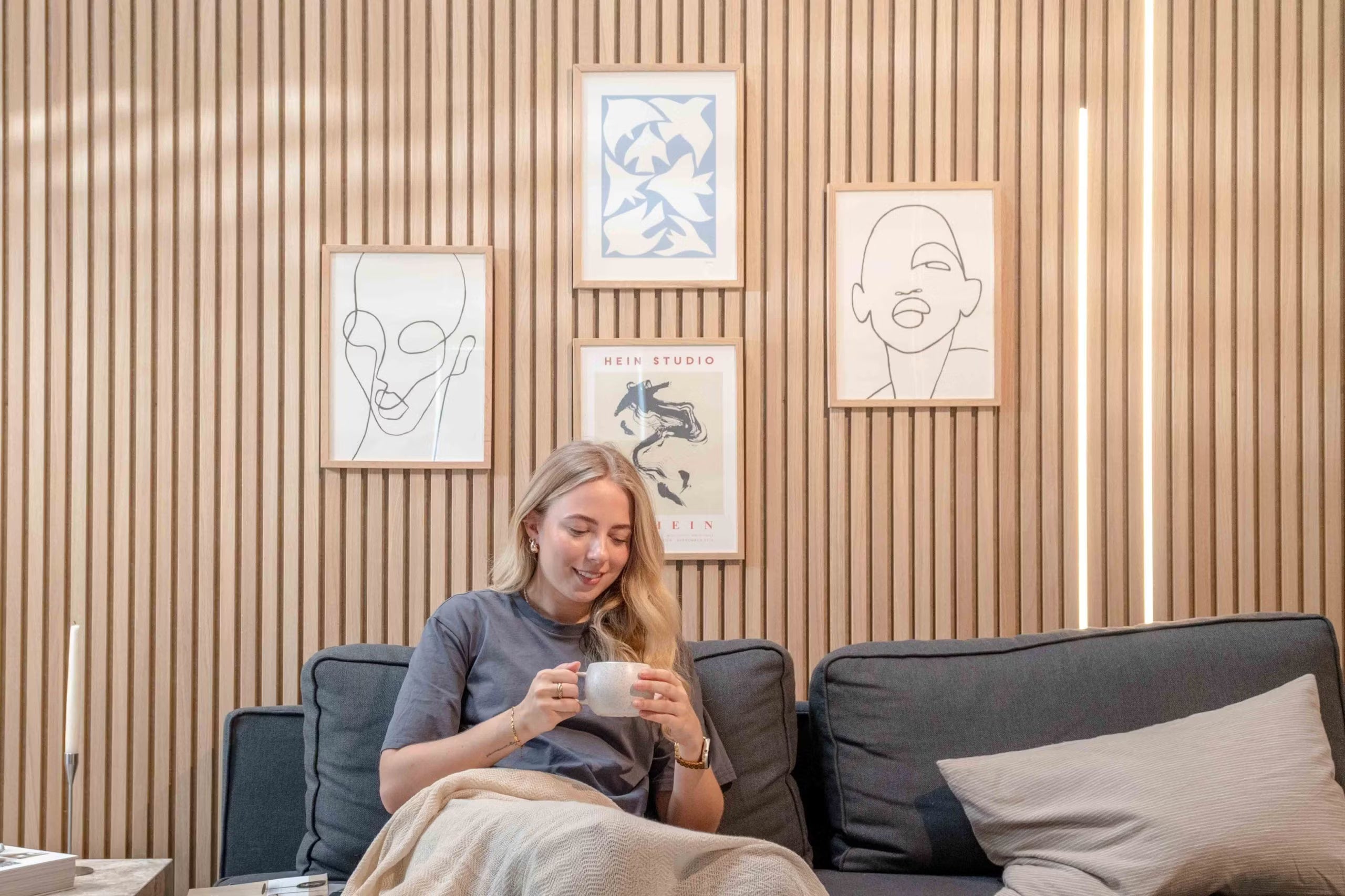 Kvinde slapper af i en sofa med et krus og smiler under minimalistisk stregkunst og abstrakt dekoration monteret på teksturerede nordiske akustiske lamelvægspaneler.