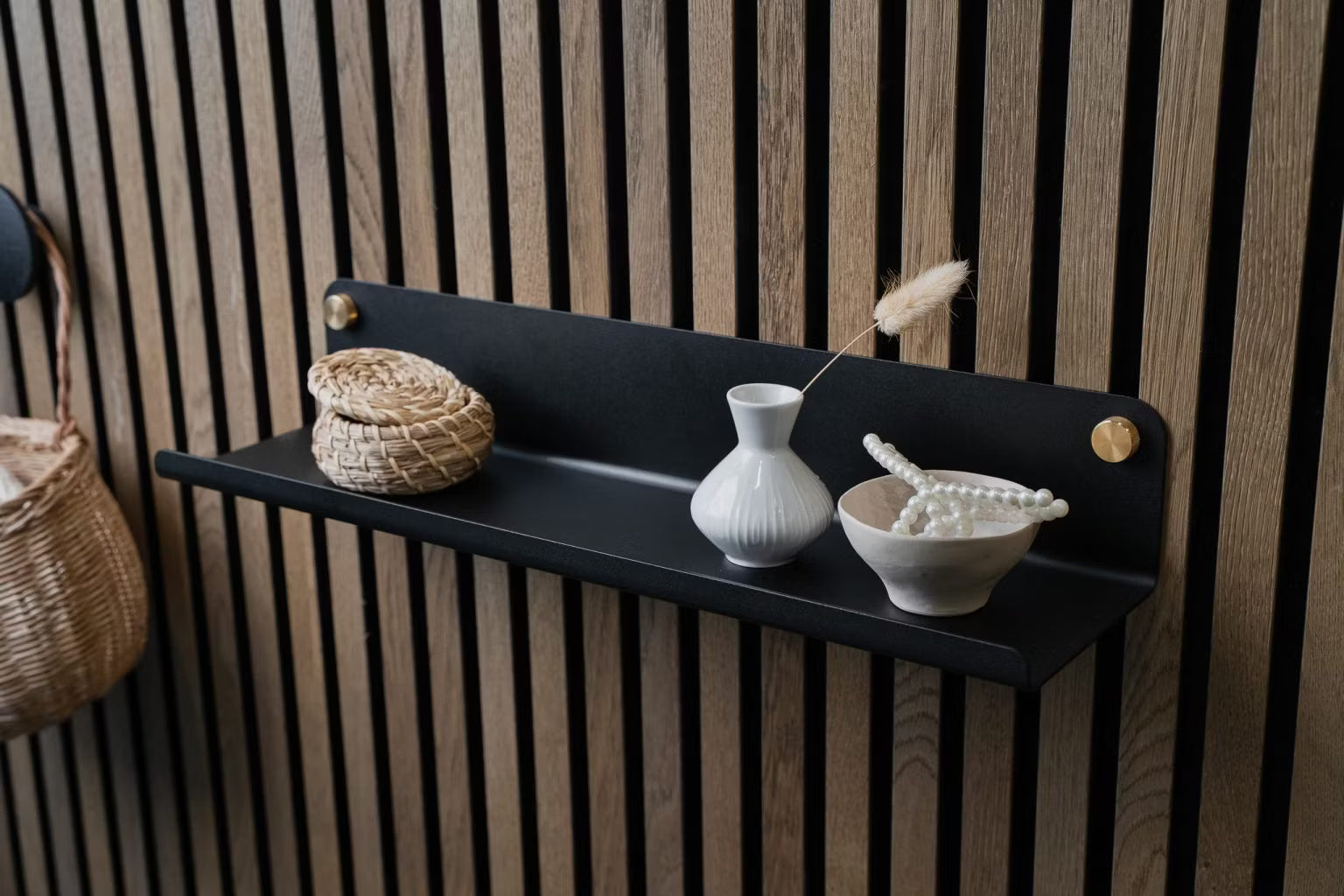 En sort væghylde med en lille flettet kurv, en hvid vase og en keramikskål med perler monteret på et nordisk akustikpanel af træ.